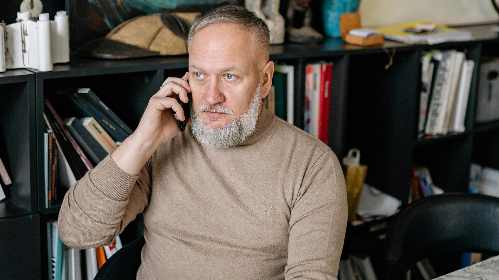 Senior man with beard conversing on phone in an art-filled workspace with modern decor.