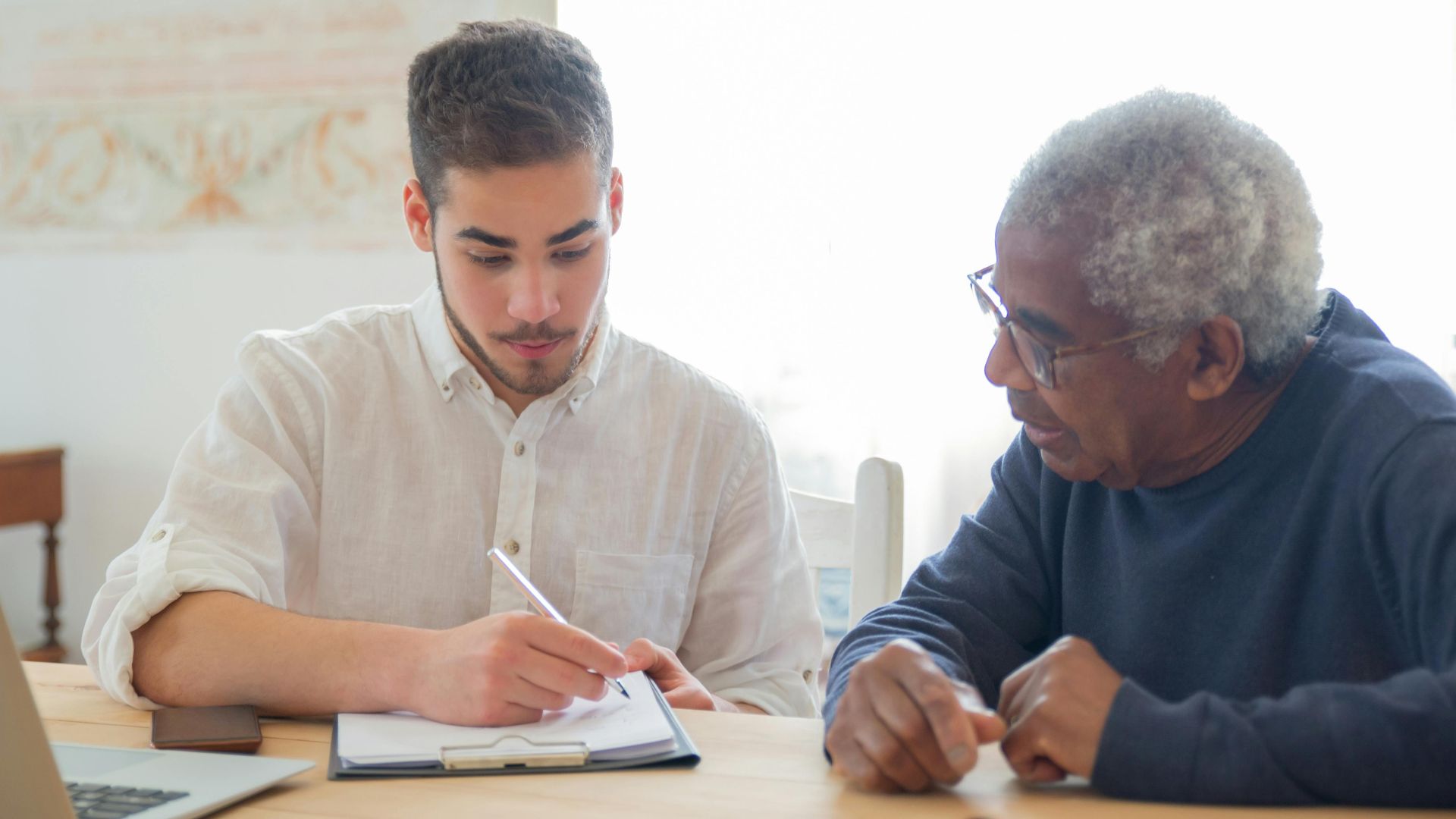 Young man helps elderly man with documents at a desk indoors, promoting collaboration.
