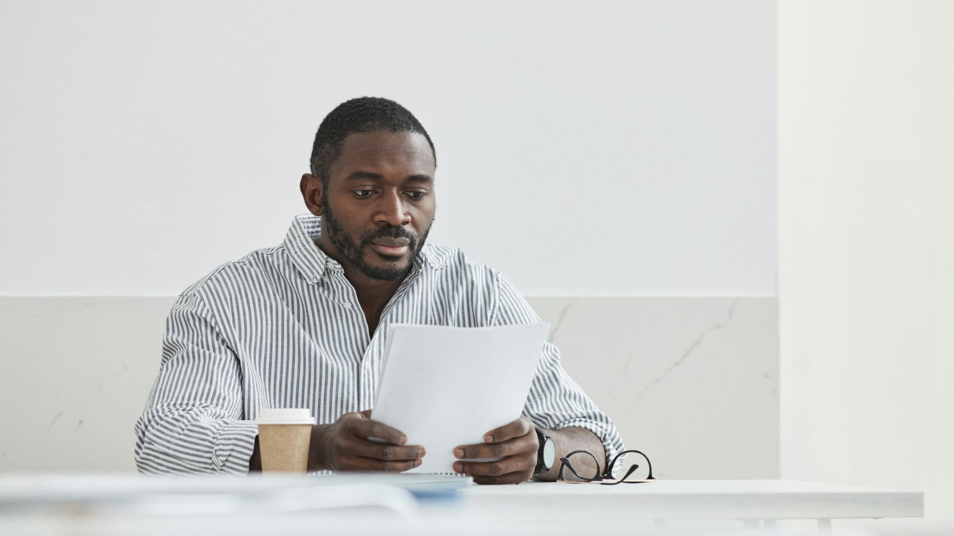 African American man sitting indoors, reading papers with a coffee cup nearby.
