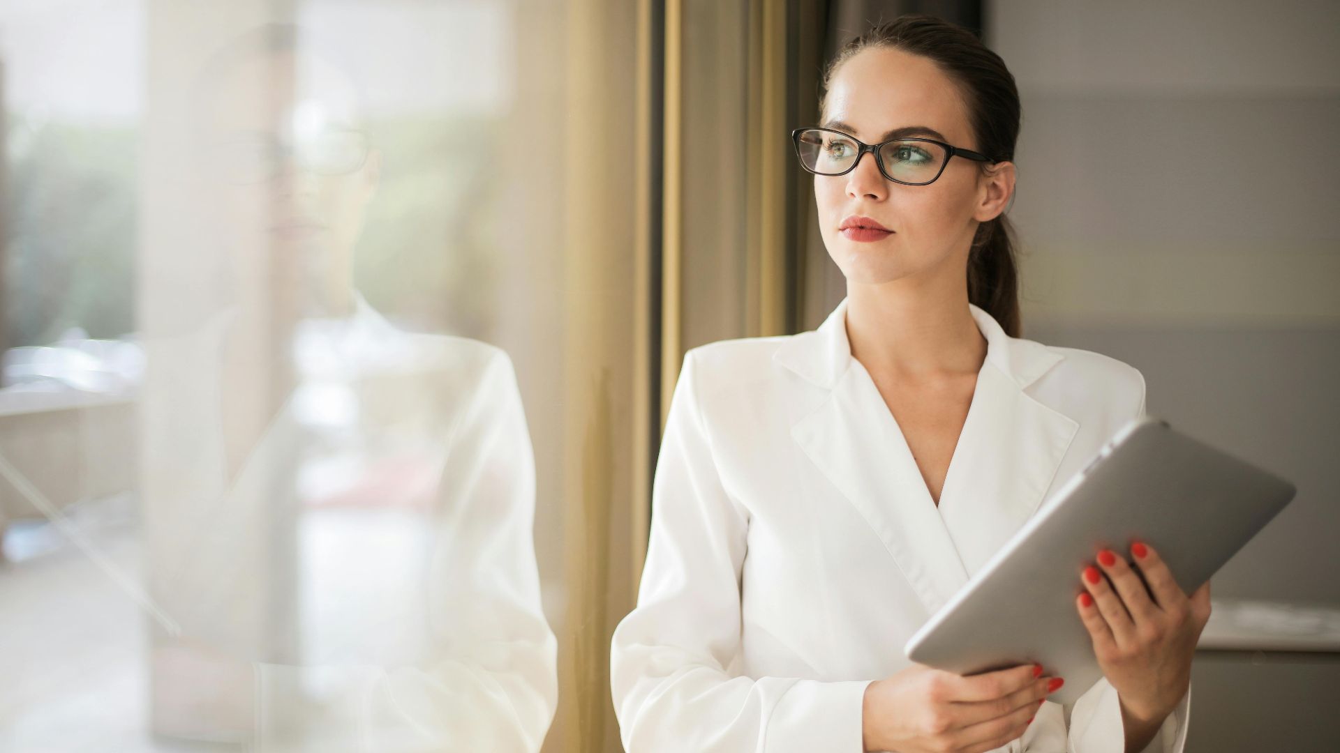 Businesswoman in formal attire thinking beside a window with a tablet.