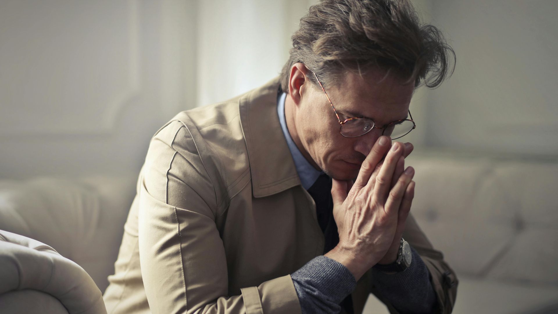 Side view of upset male entrepreneur in formal wear sitting on couch in modern living room and contemplating about financial problems while leaning on hands and looking down