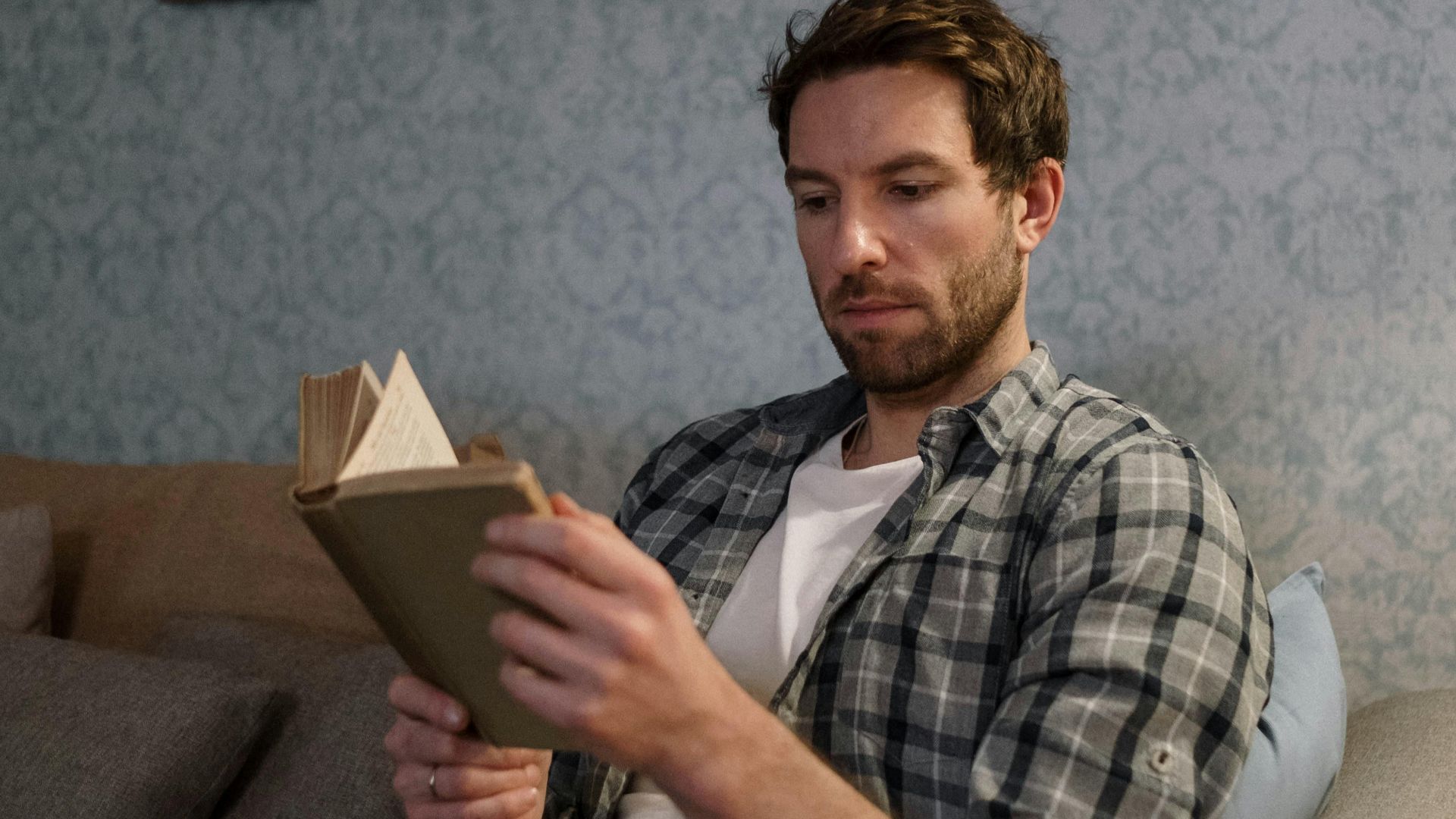 Bearded man in plaid shirt reading on a cozy sofa for leisure and relaxation.