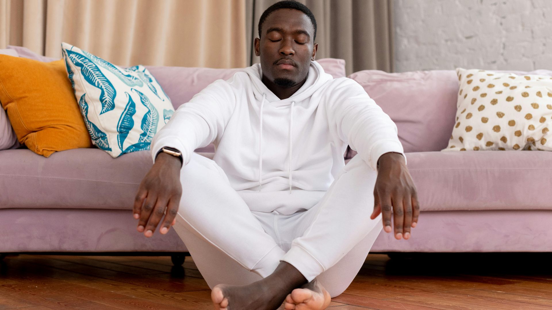 Calm African American male in white clothes sitting with closed eyes and crossed legs on floor against comfortable sofa in living room