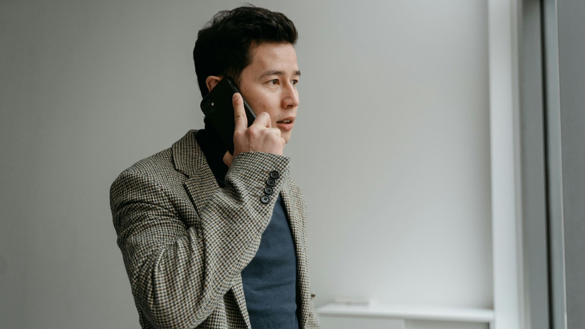 Young businessman in blazer talking on smartphone near window indoors.