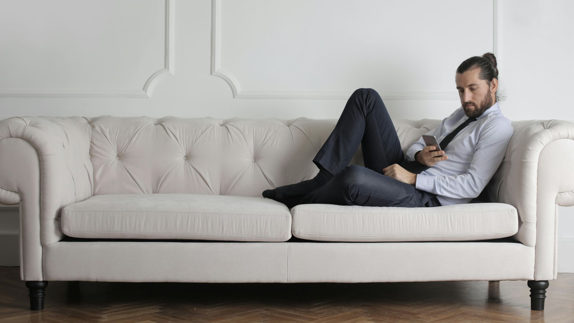 A man relaxes on a stylish white sofa in a minimalist living room, using a smartphone.