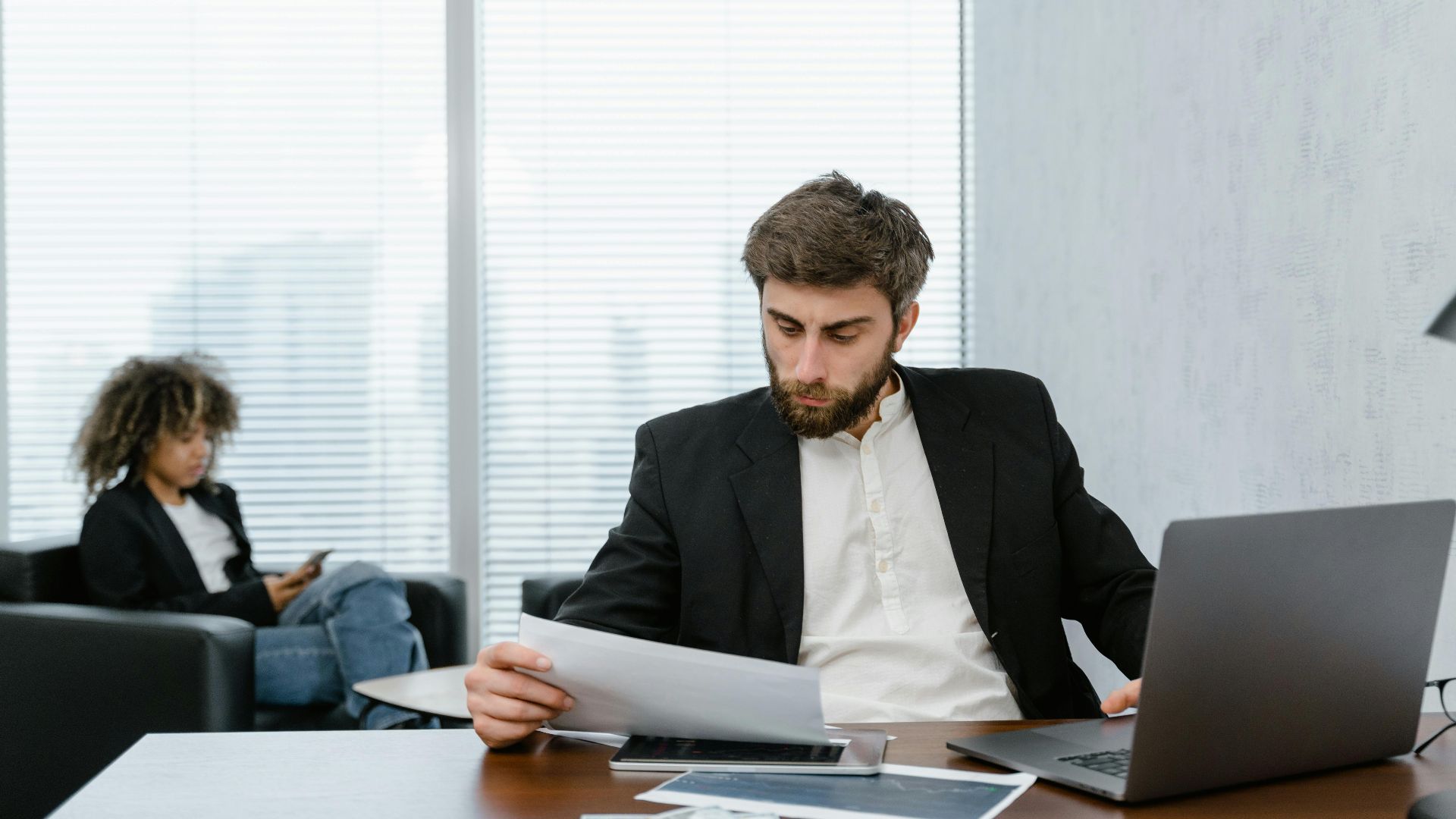 Focused businessman reviews financial reports at office desk with laptop.