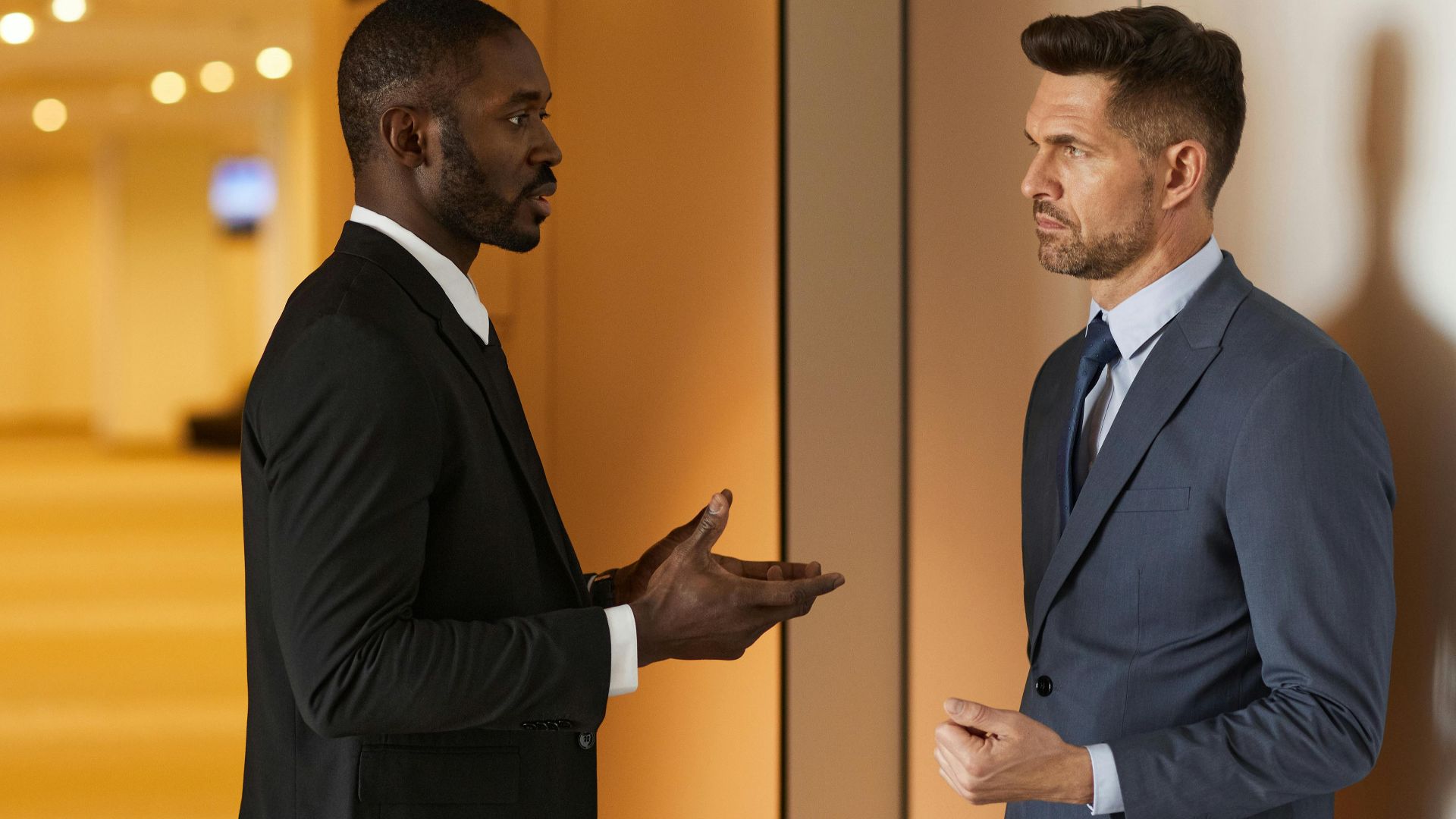 Two men in suits having a discussion in a modern hallway setting.