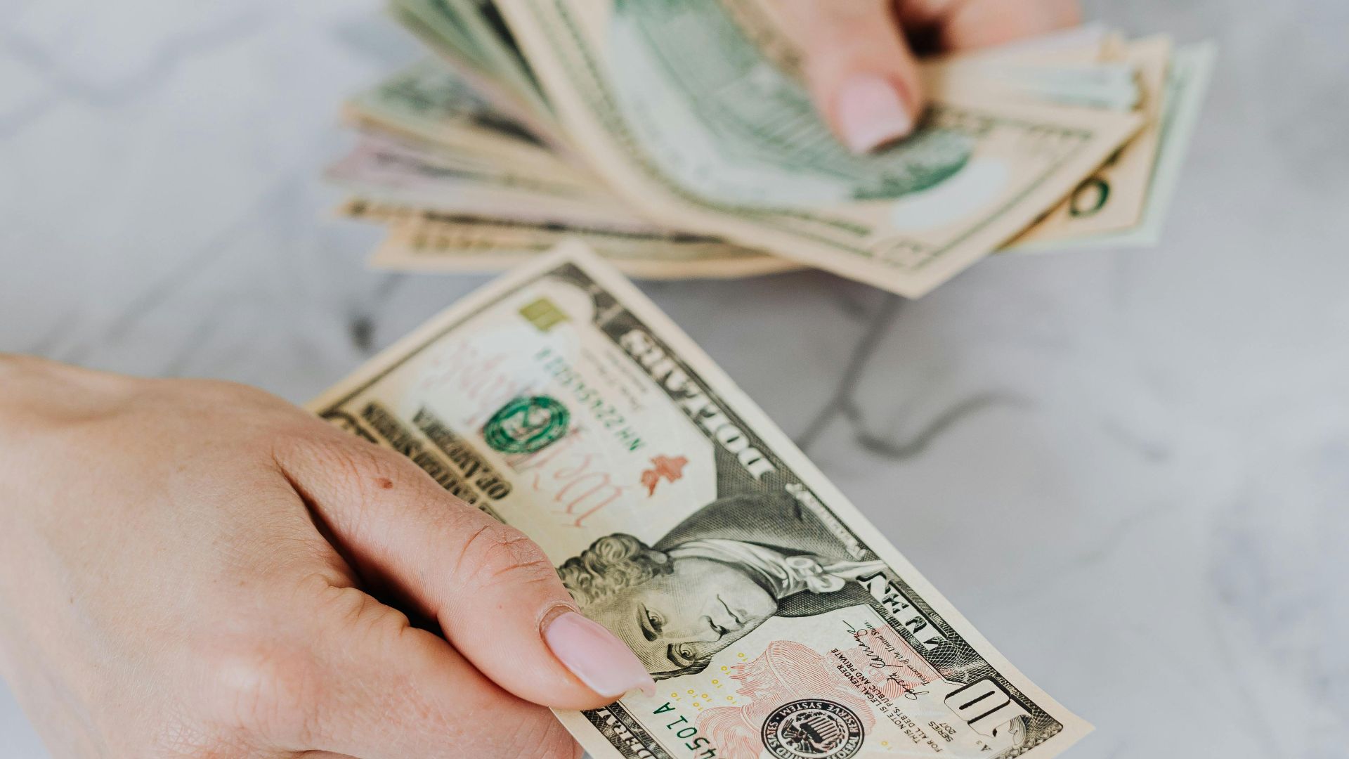 Close-up of hands counting US dollar bills on a marble table, symbolizing personal finance.
