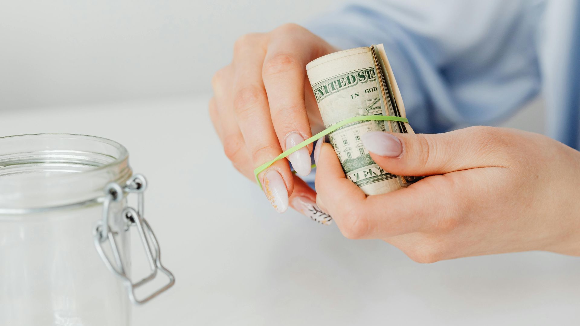 Close-up of hands securing rolled US dollar bills with rubber band beside a glass jar.