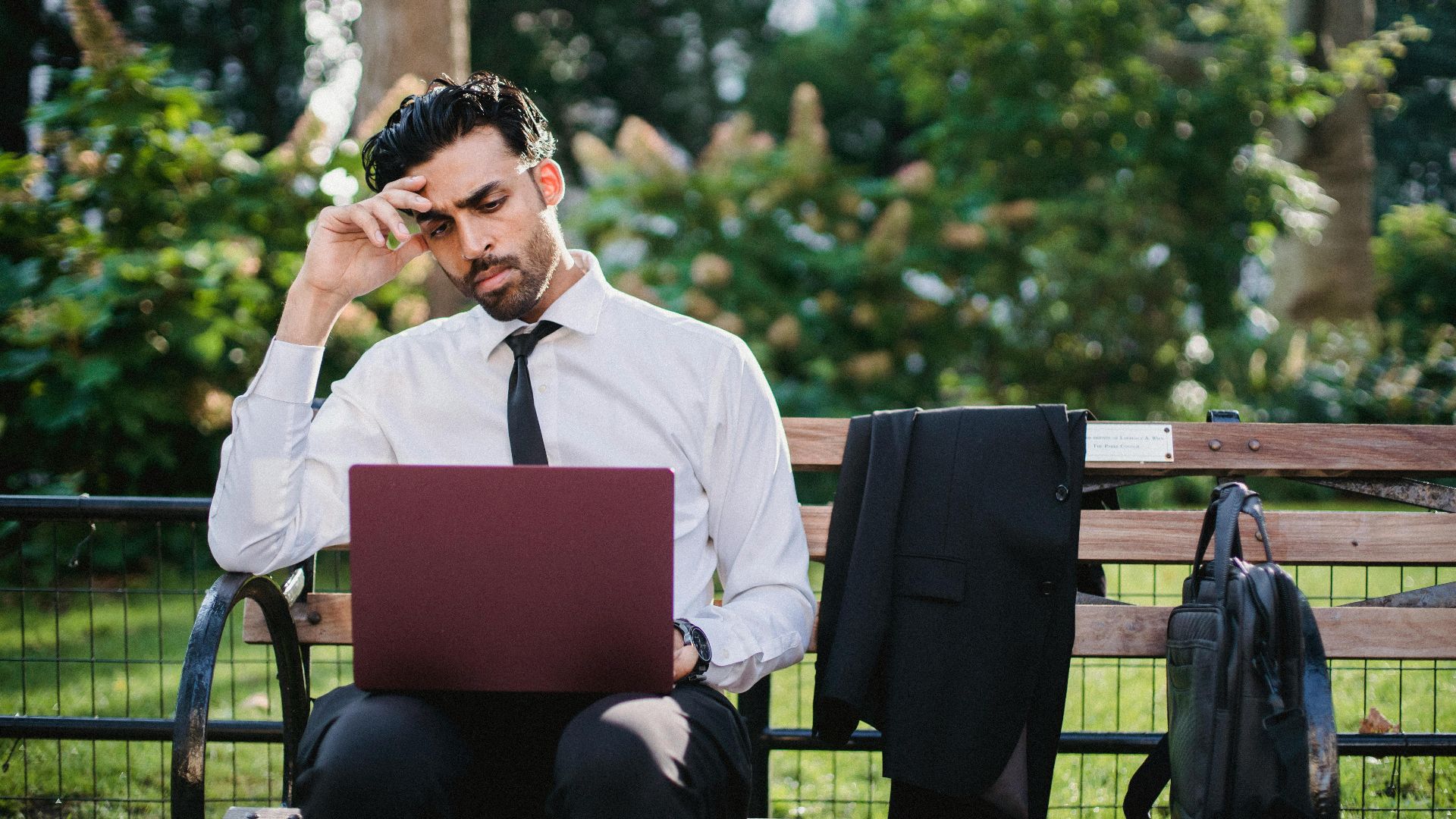 A thoughtful businessman works on his laptop while sitting on a park bench with his coat and bag.