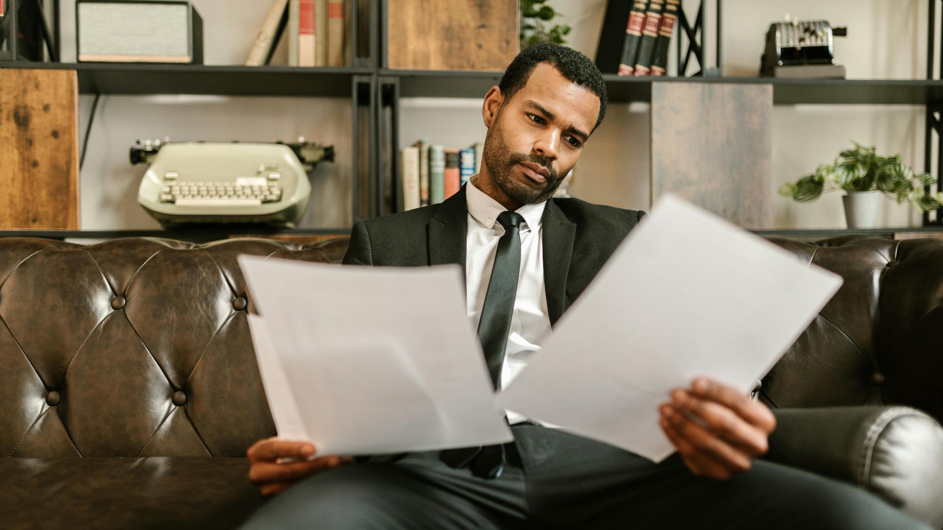 A professional businessman intently reviews documents while seated on a leather sofa in a modern office setting.