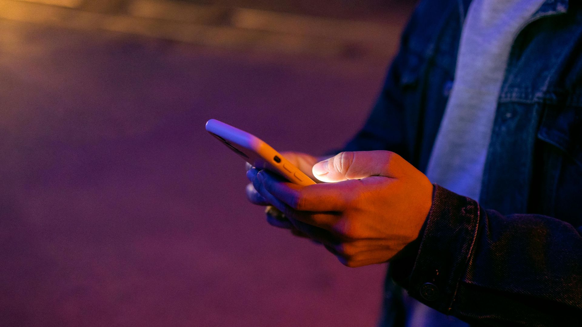 A person holds a smartphone outdoors at night, illuminated by colorful ambient lights.
