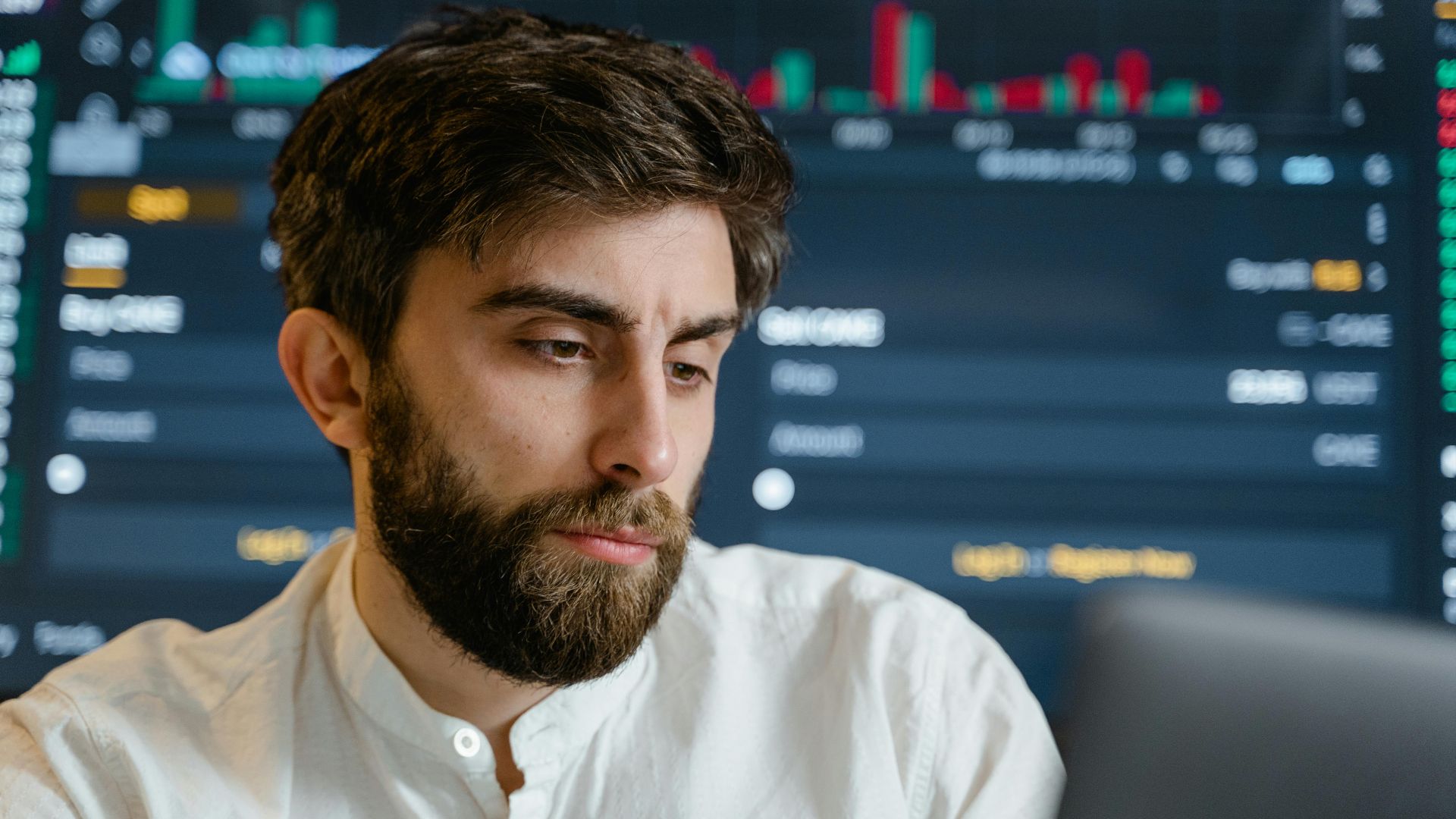 Close-up of thoughtful businessman with beard analyzing stock market data on screen.