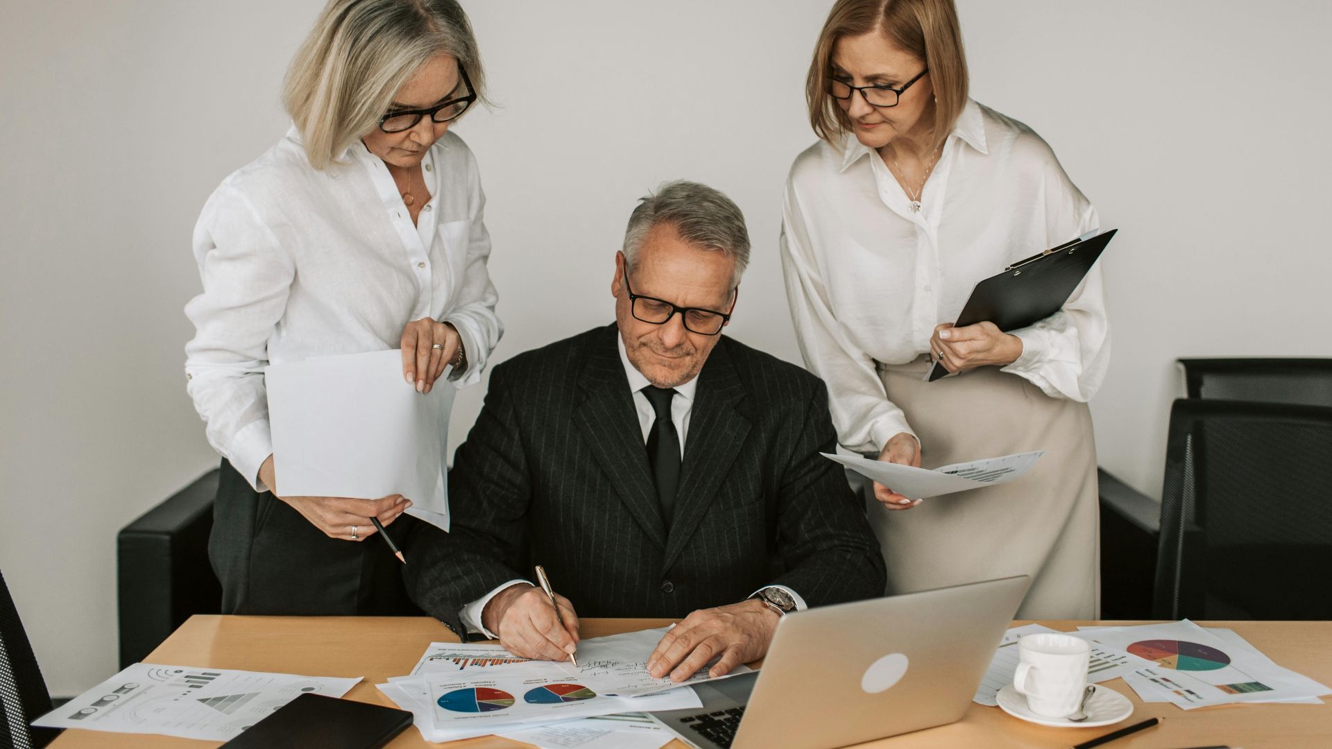 Three mature professionals collaborate at an office desk, analyzing charts and documents.