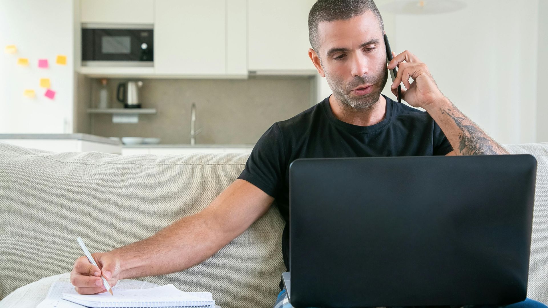 Man working from home using a laptop and smartphone while taking notes in a modern kitchen setting.