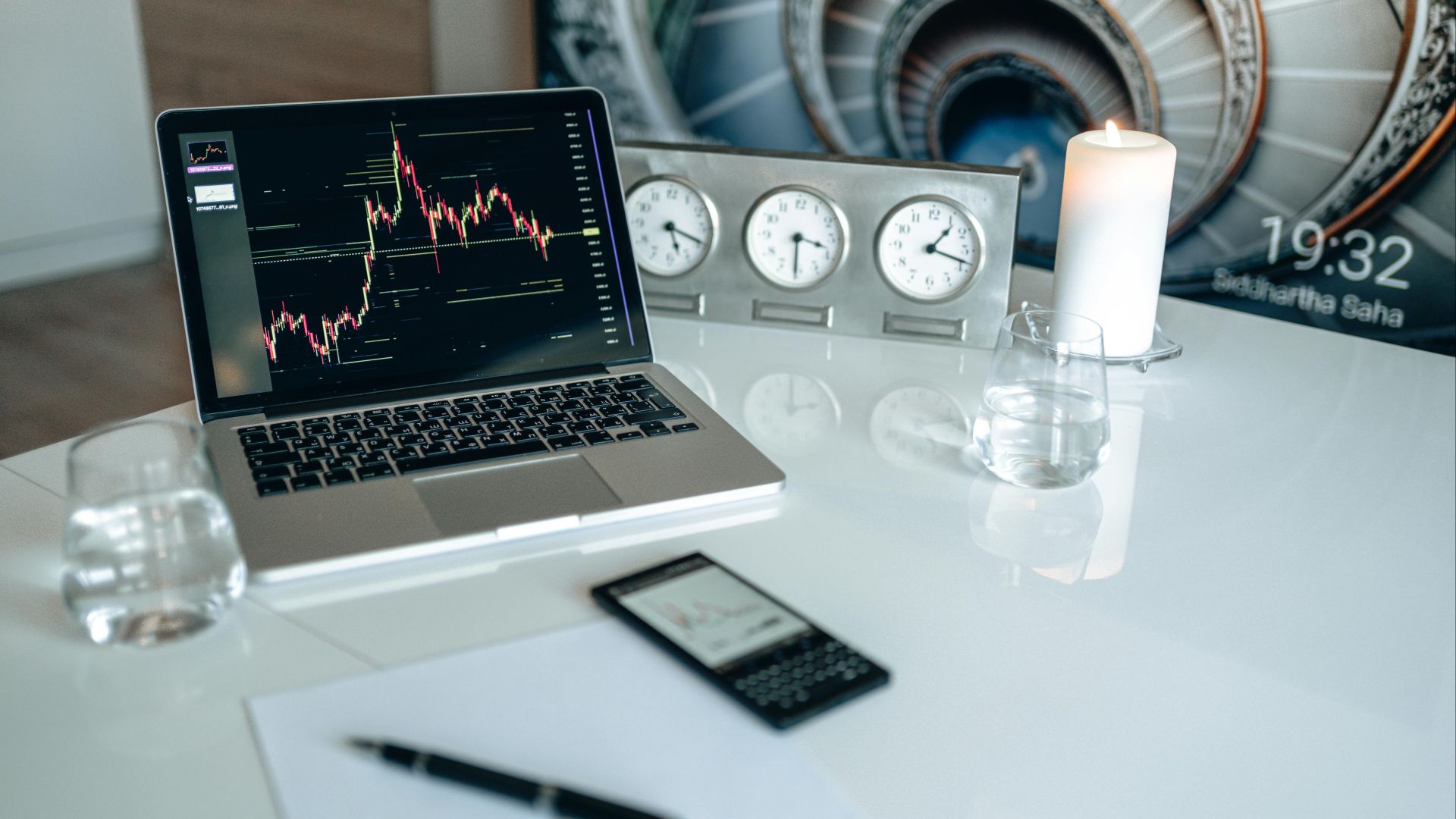 A modern desk setup featuring a laptop displaying trading charts, candles, and clocks.