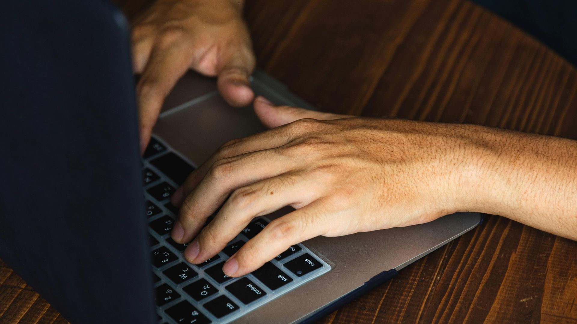 Close-up of a man typing on a laptop at a wooden table, indoor setting.