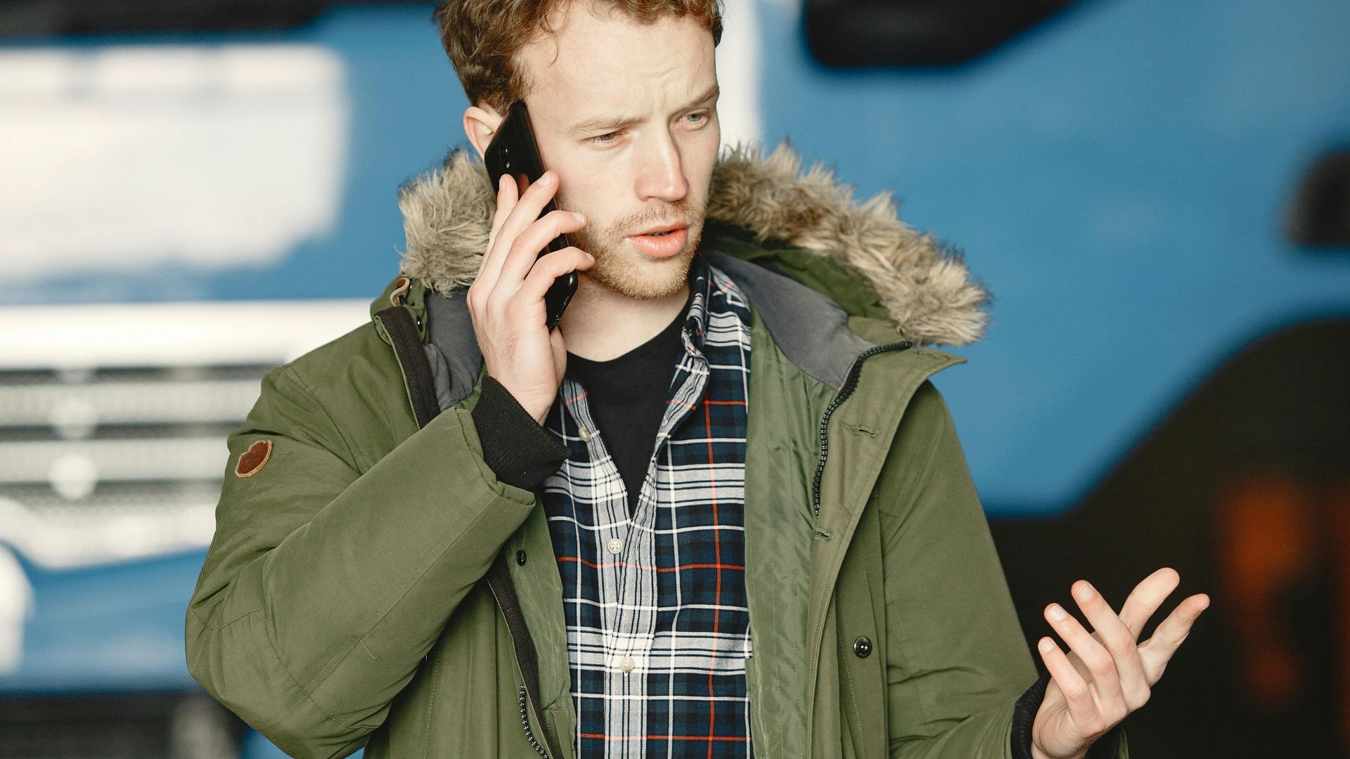 Man wearing green jacket and plaid shirt talks on the phone near a truck outdoors.