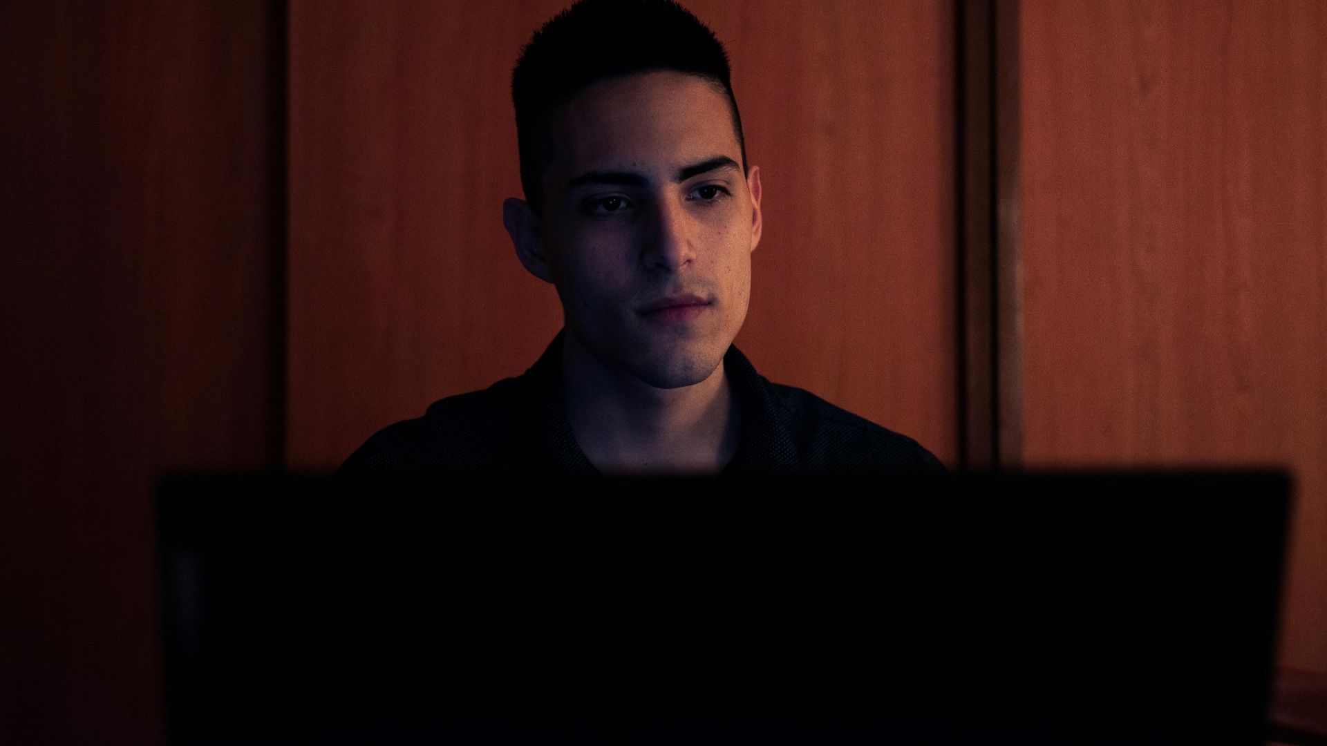 A young man working on his laptop, focused and serious, in an indoor setting with warm lighting.