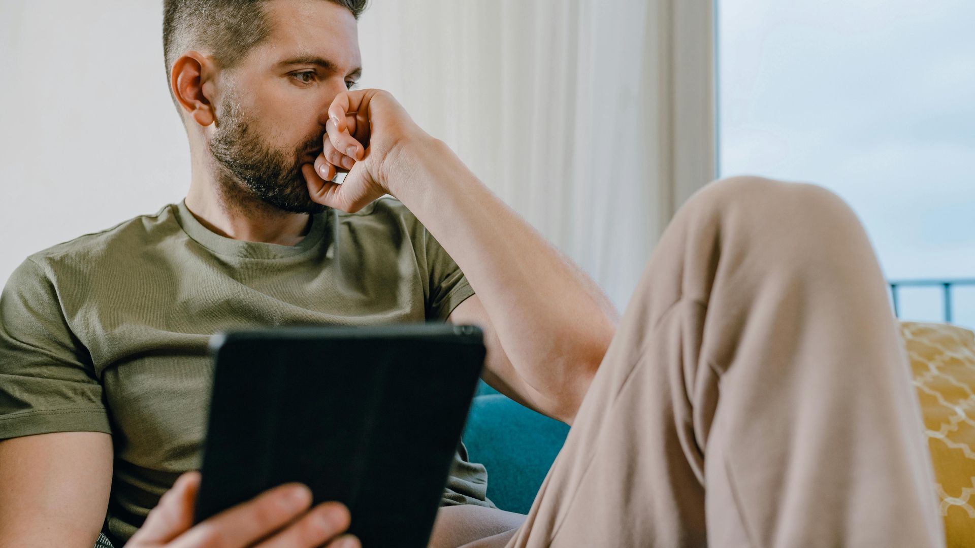 A man in casual attire, sitting indoors, holding a tablet and looking thoughtful.