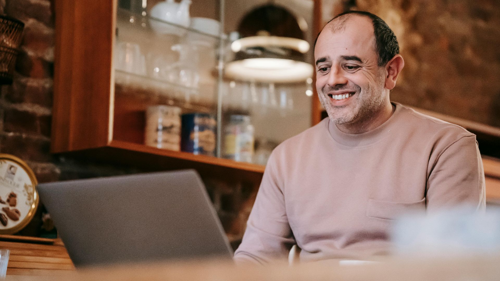 Content adult ethnic male freelancer in casual clothes smiling while working online on laptop sitting at table at home