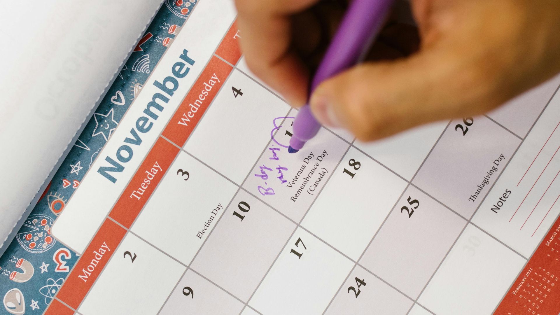 A person writes a reminder on a colorful November calendar using a purple pen.