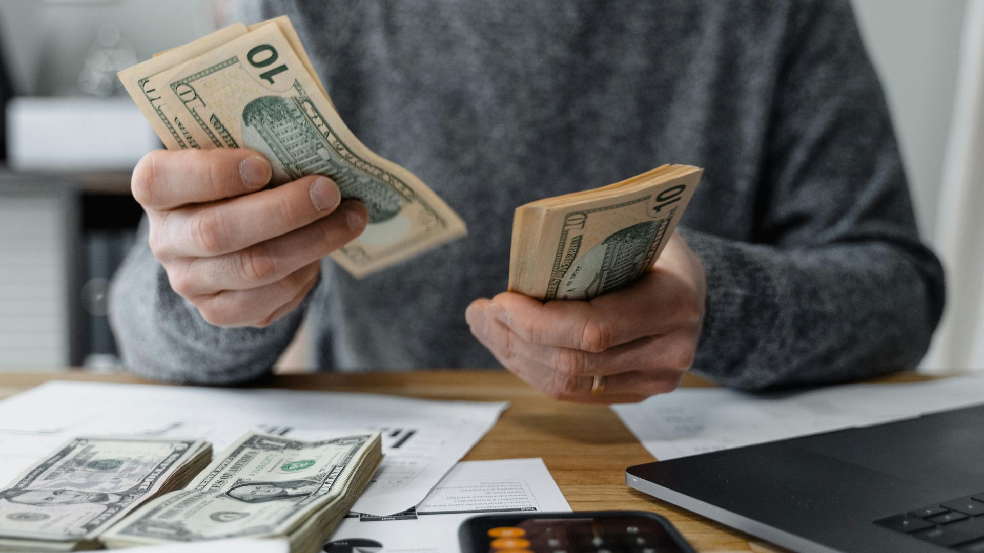 Close-up of person counting cash with a calculator and paperwork on a desk.