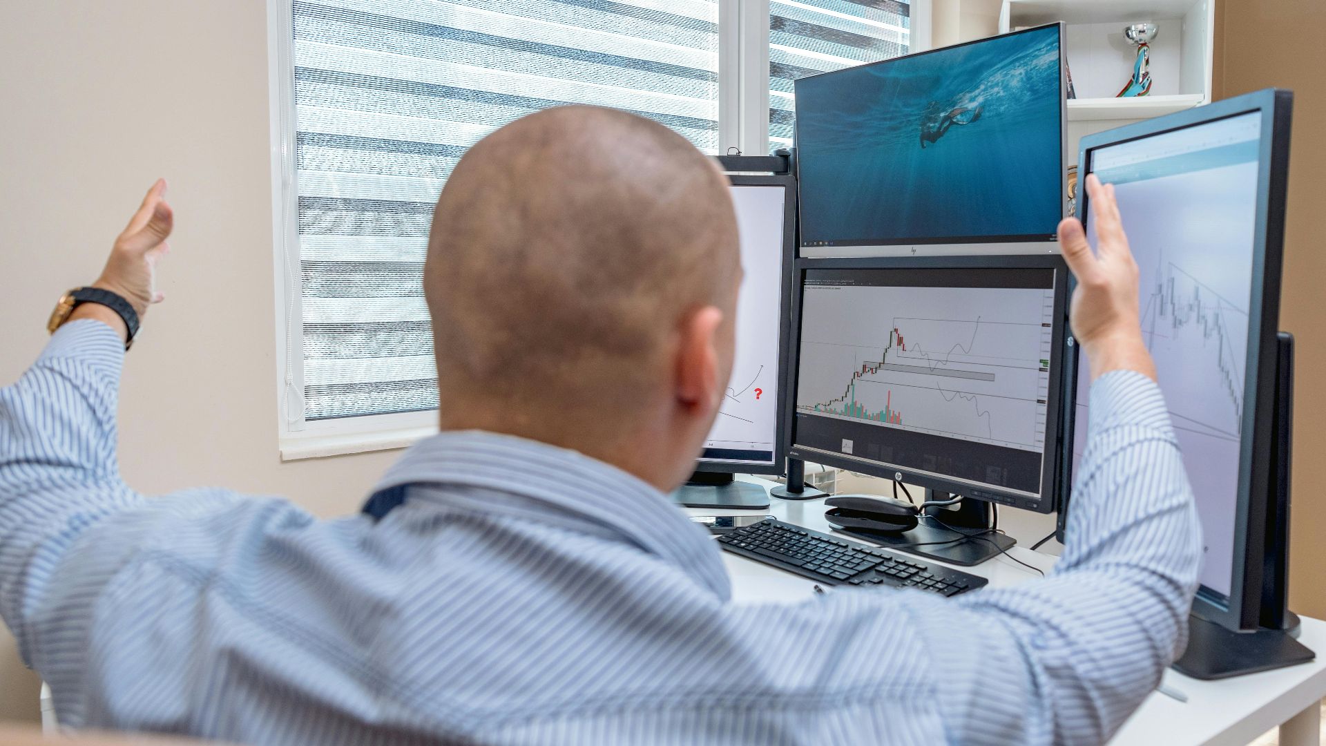 A stock trader in an office raises his hands in celebration while monitoring multiple screens with financial charts.