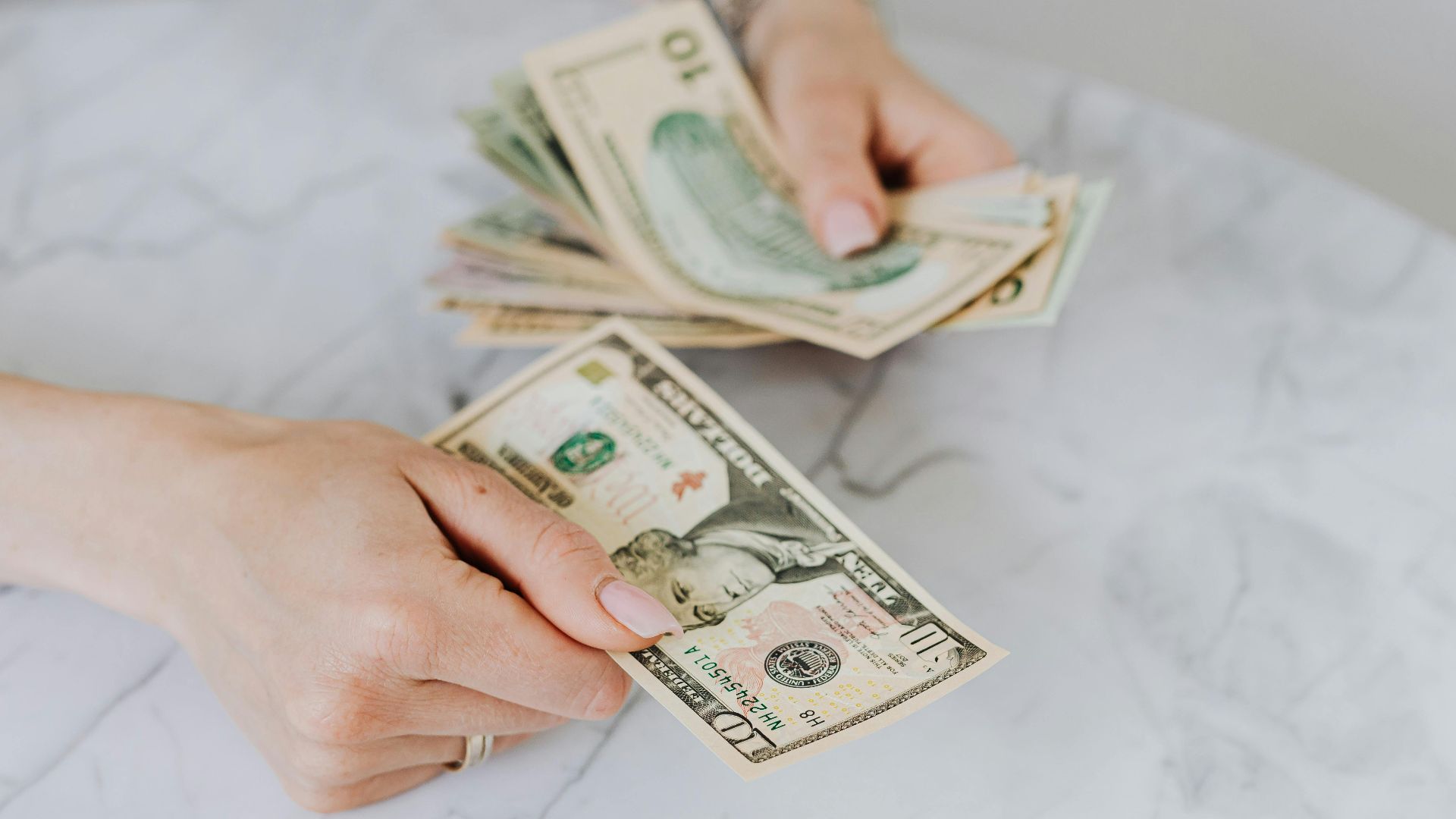 Close-up of hands counting US dollar bills on a marble table surface.
