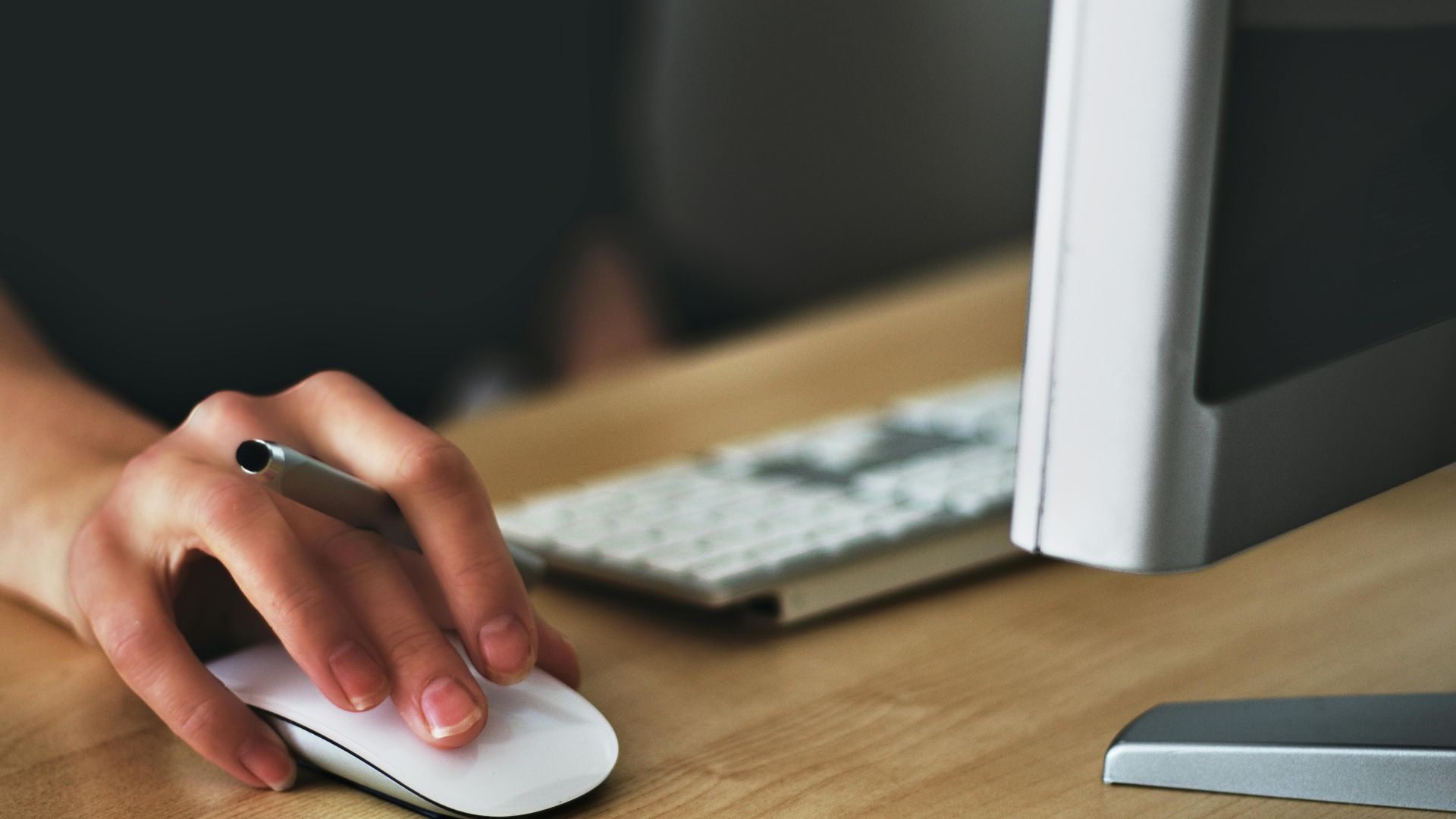 A hand using a wireless mouse at a modern desk setup with a computer and keyboard.