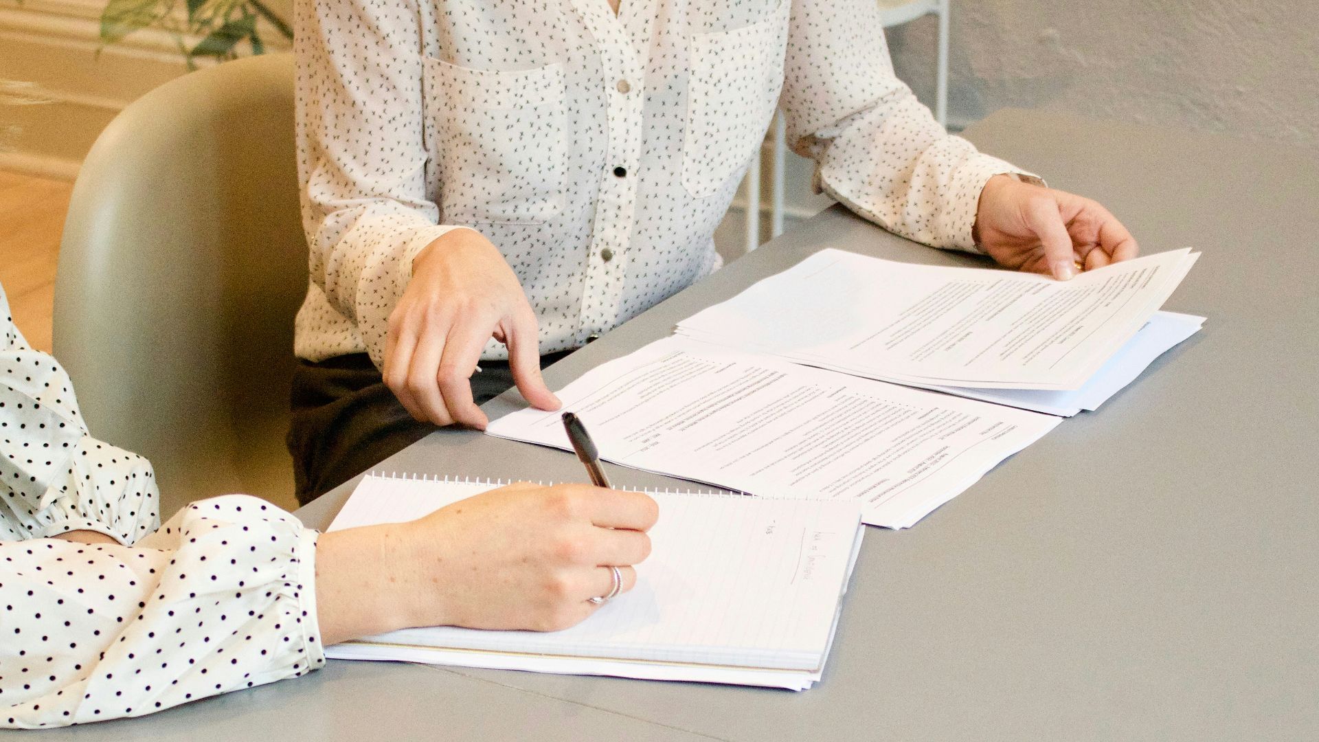 woman signing on white printer paper beside woman about to touch the documents
