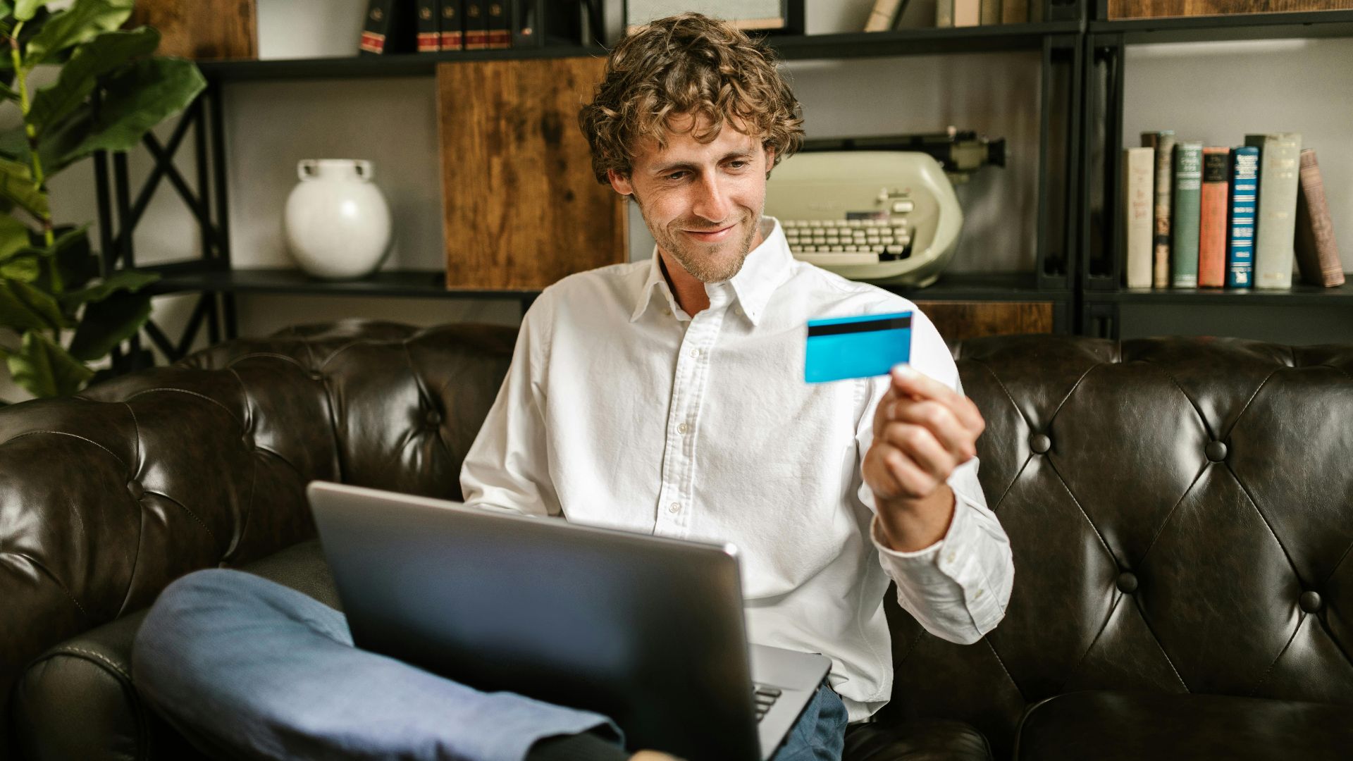 A man sits on a sofa, smiling while holding a credit card with a laptop on his lap, indoors.