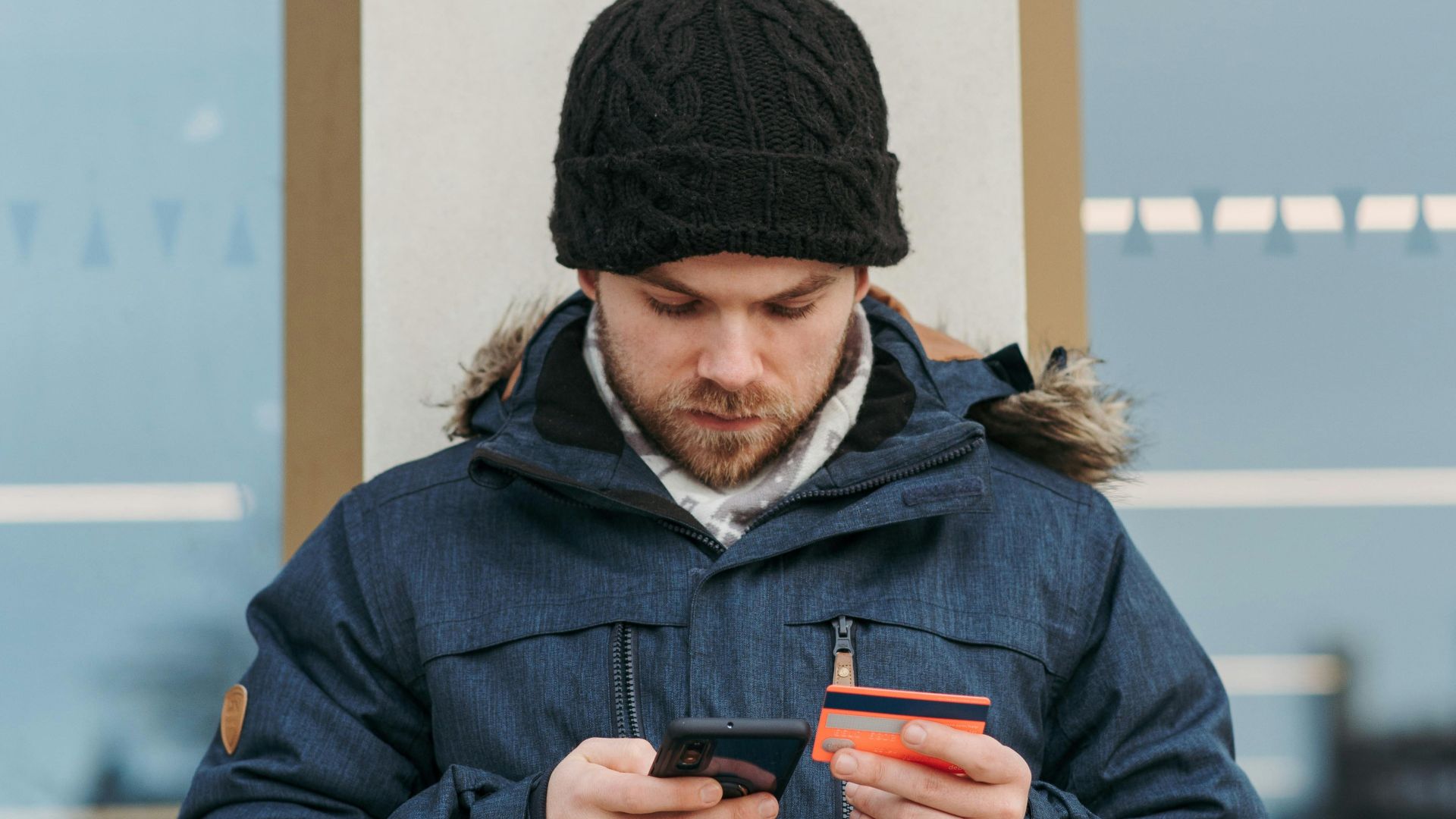Serious young guy in warm clothes and hat standing on city street and browsing smartphone while making online payment with credit card near modern building