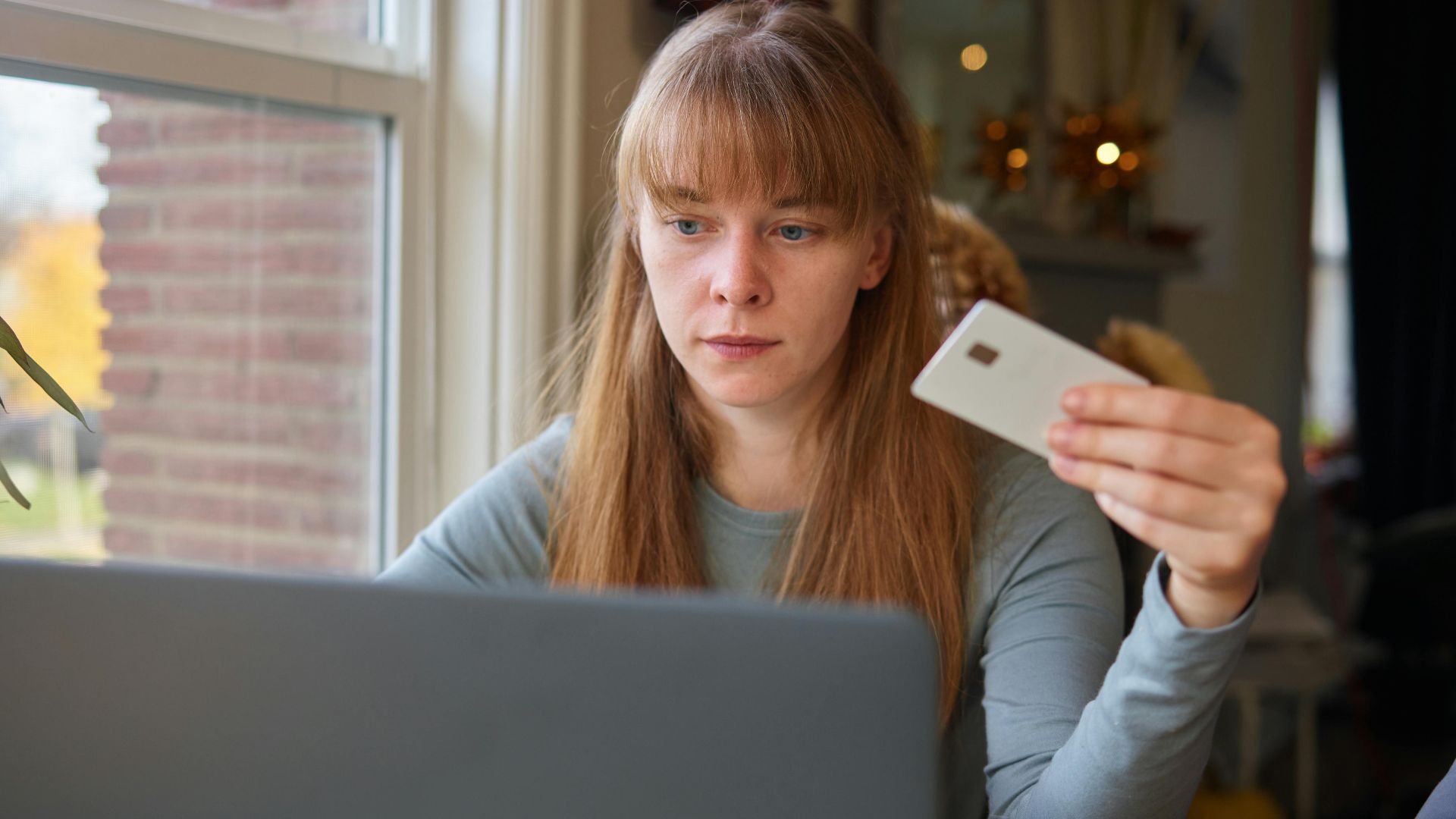 A young woman holds a credit card while using her laptop indoors.