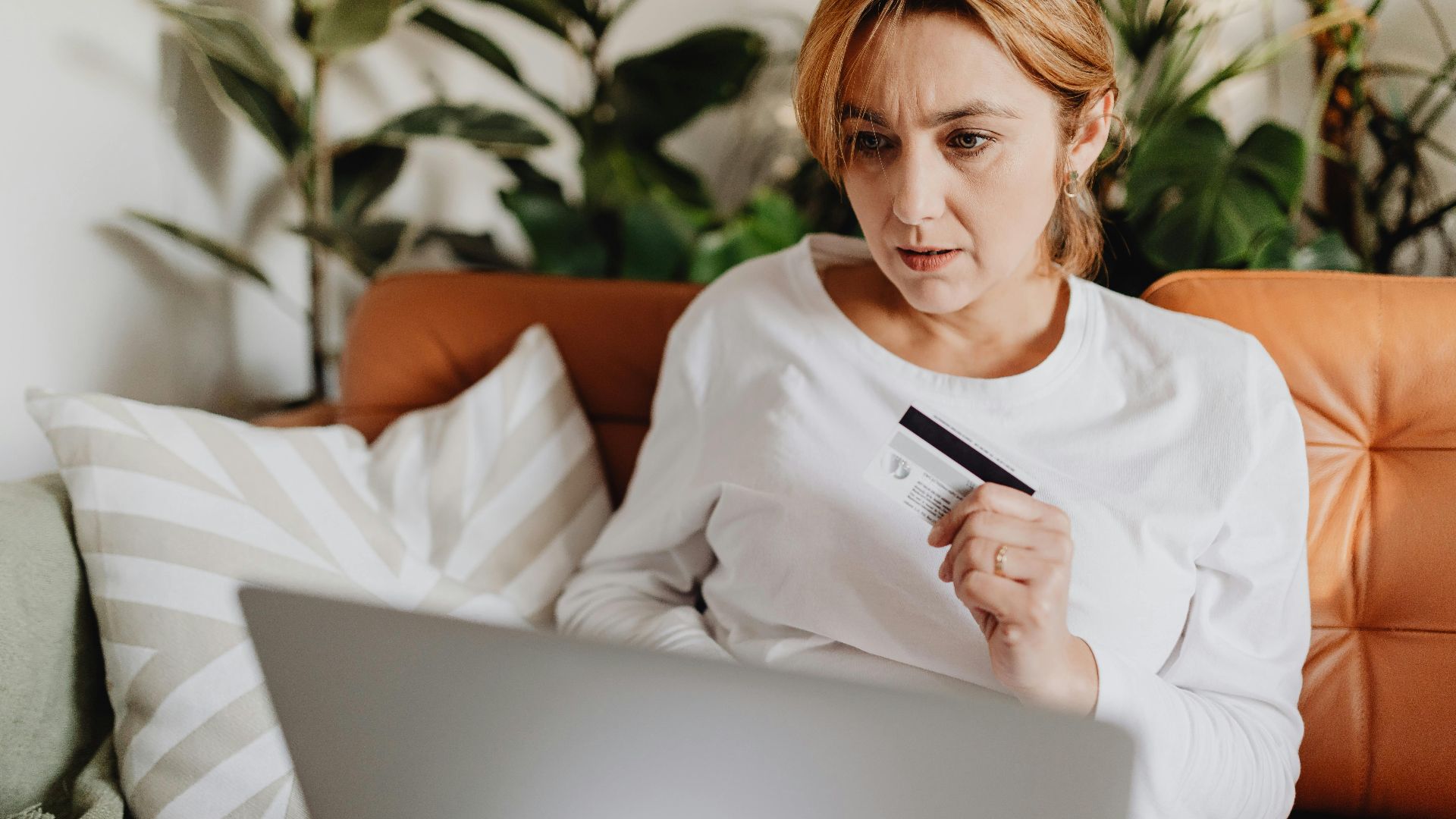 A woman sitting on a couch shopping online with a credit card and laptop in a cozy living room.