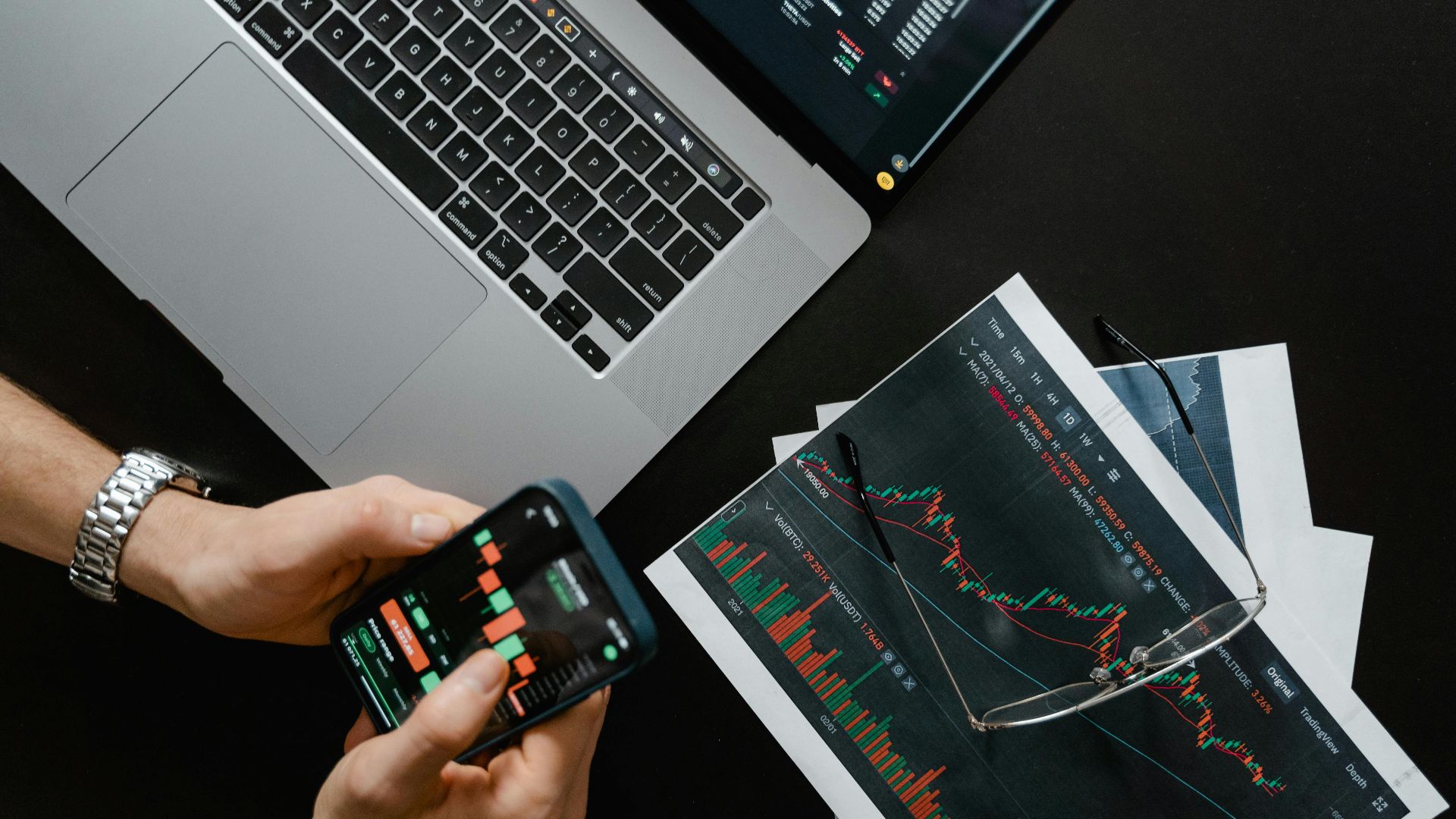 Hands using smartphone beside laptop with stock charts, showcasing digital trading.