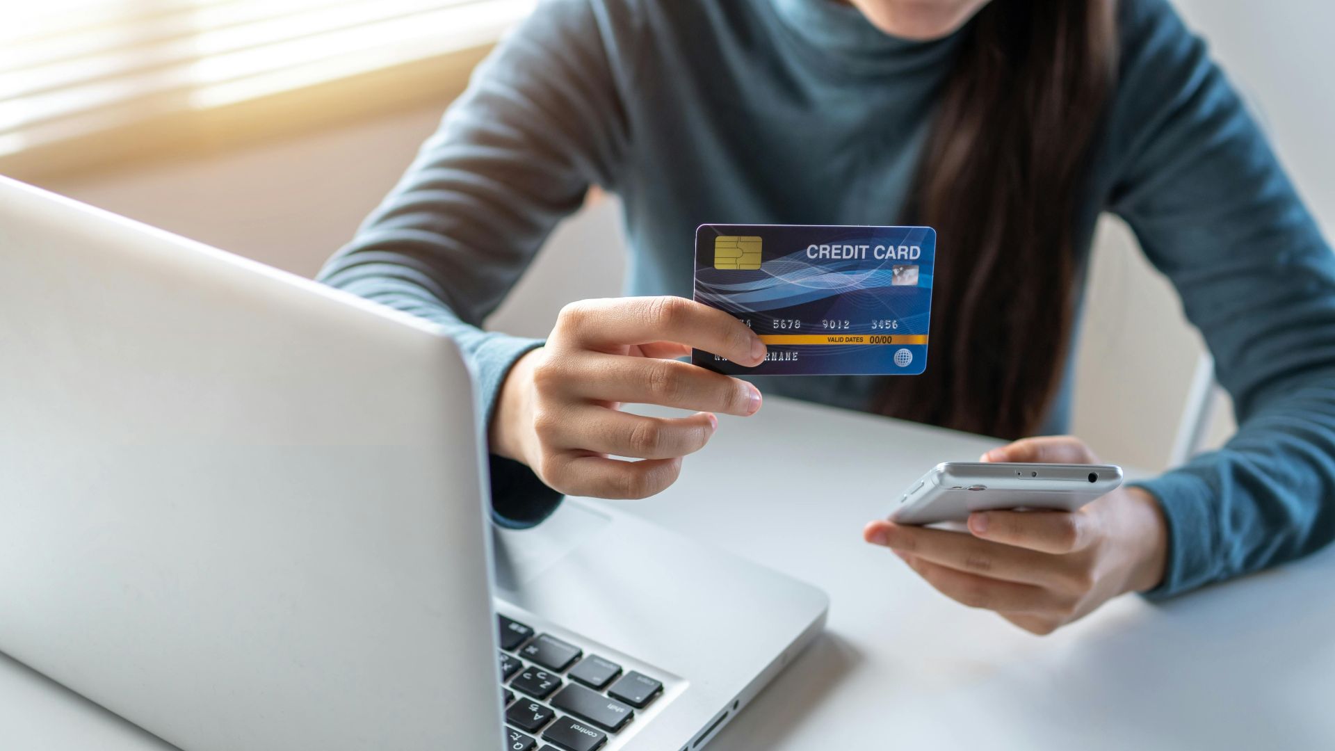 A woman uses a credit card and smartphone for online shopping on her laptop