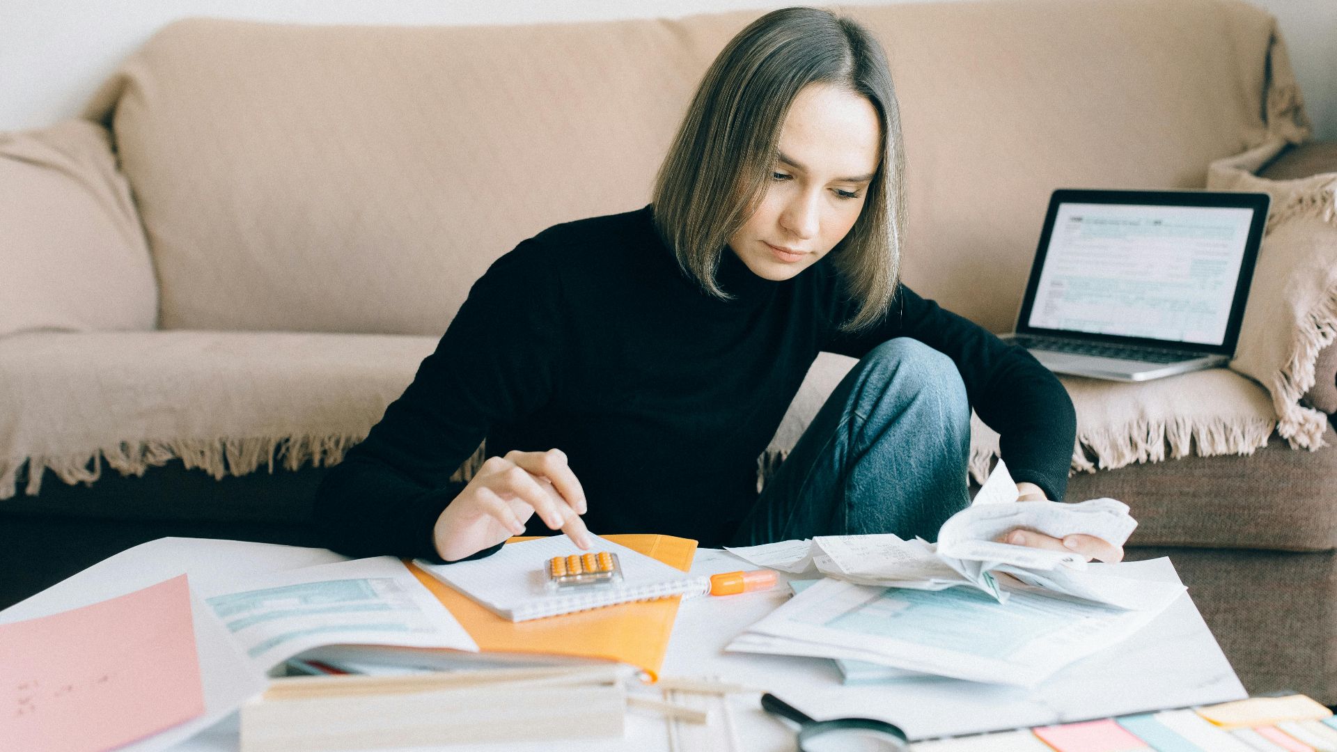 Young woman handling financial tasks with papers and laptop in cozy living room.