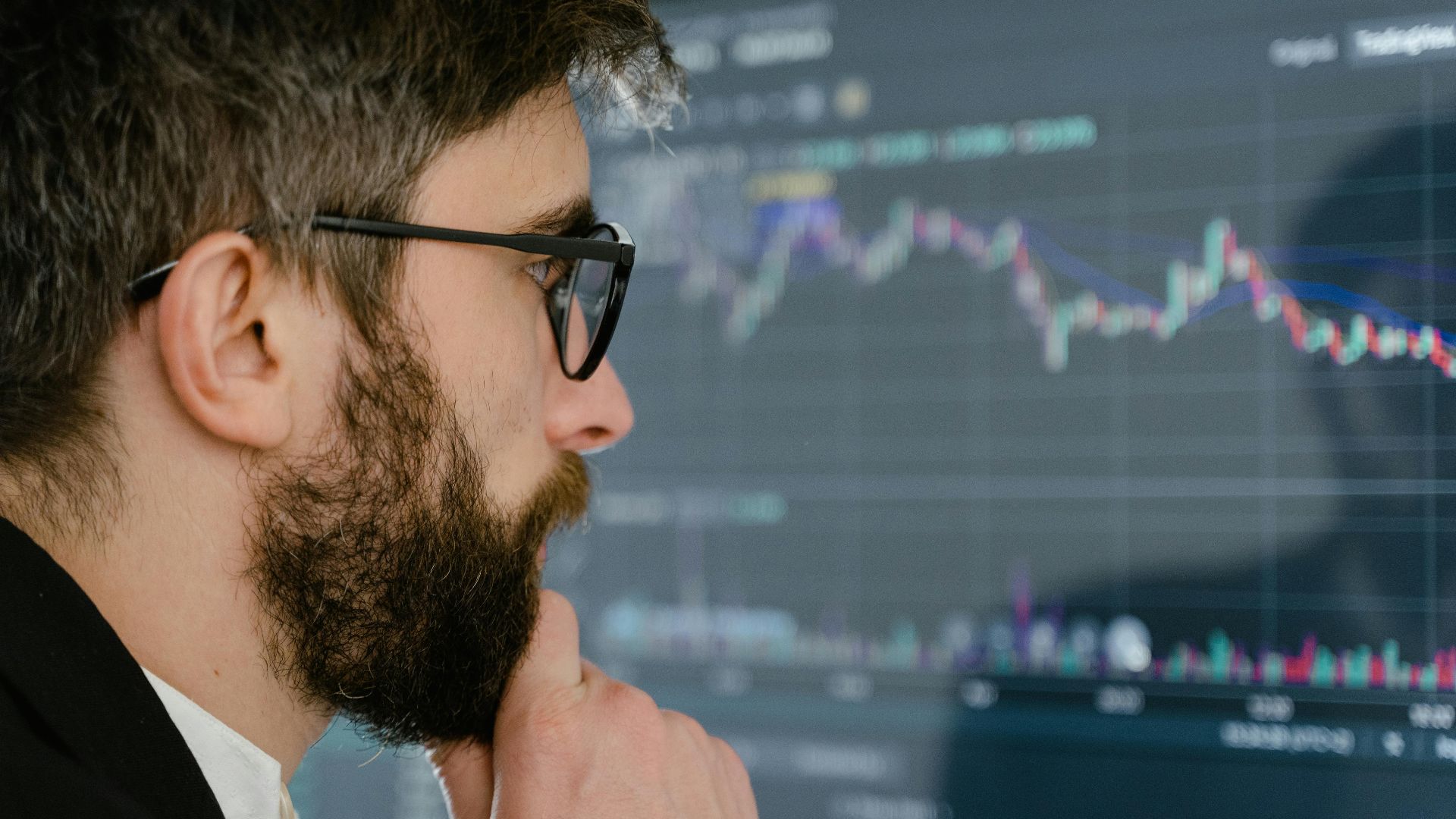 A focused man with glasses studies stock market graphs on a screen, pondering insights.