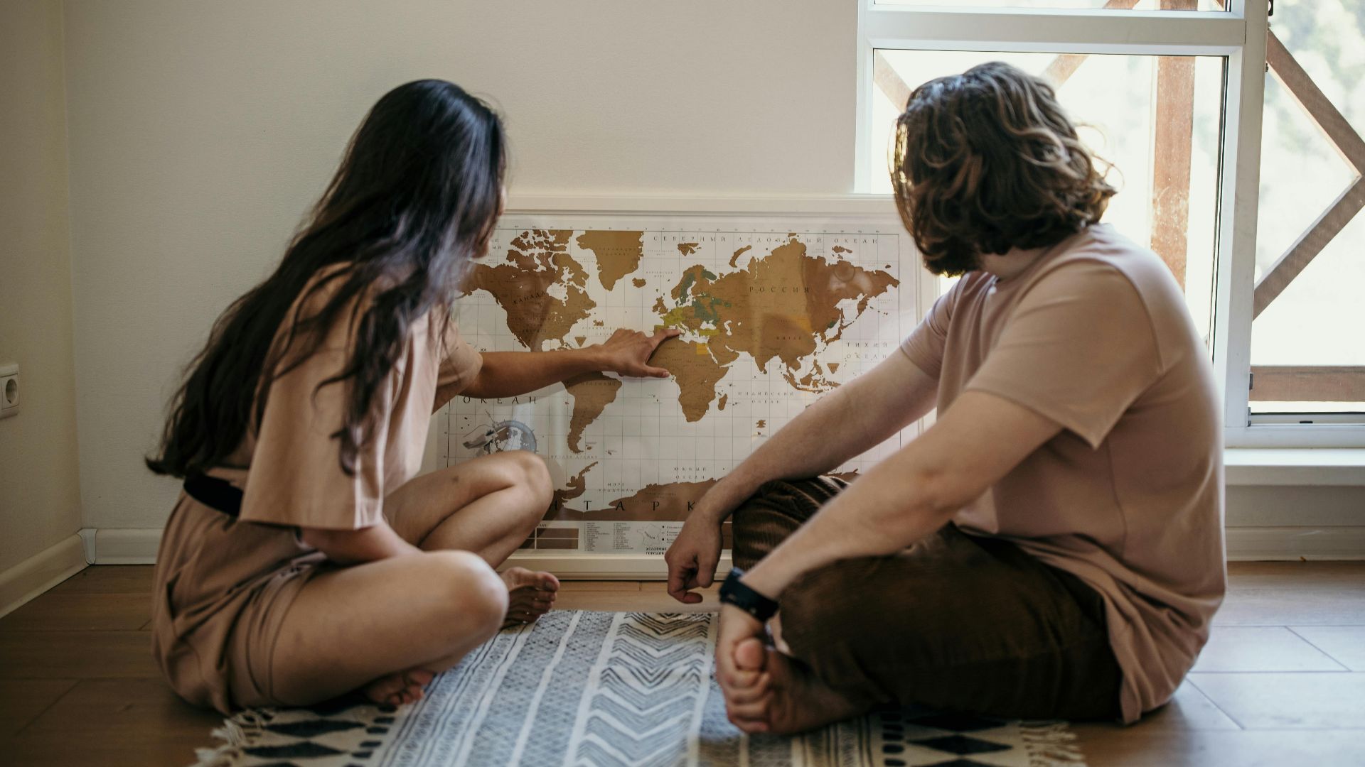 Couple sitting on floor pointing at a world map, planning their next travel adventure indoors.
