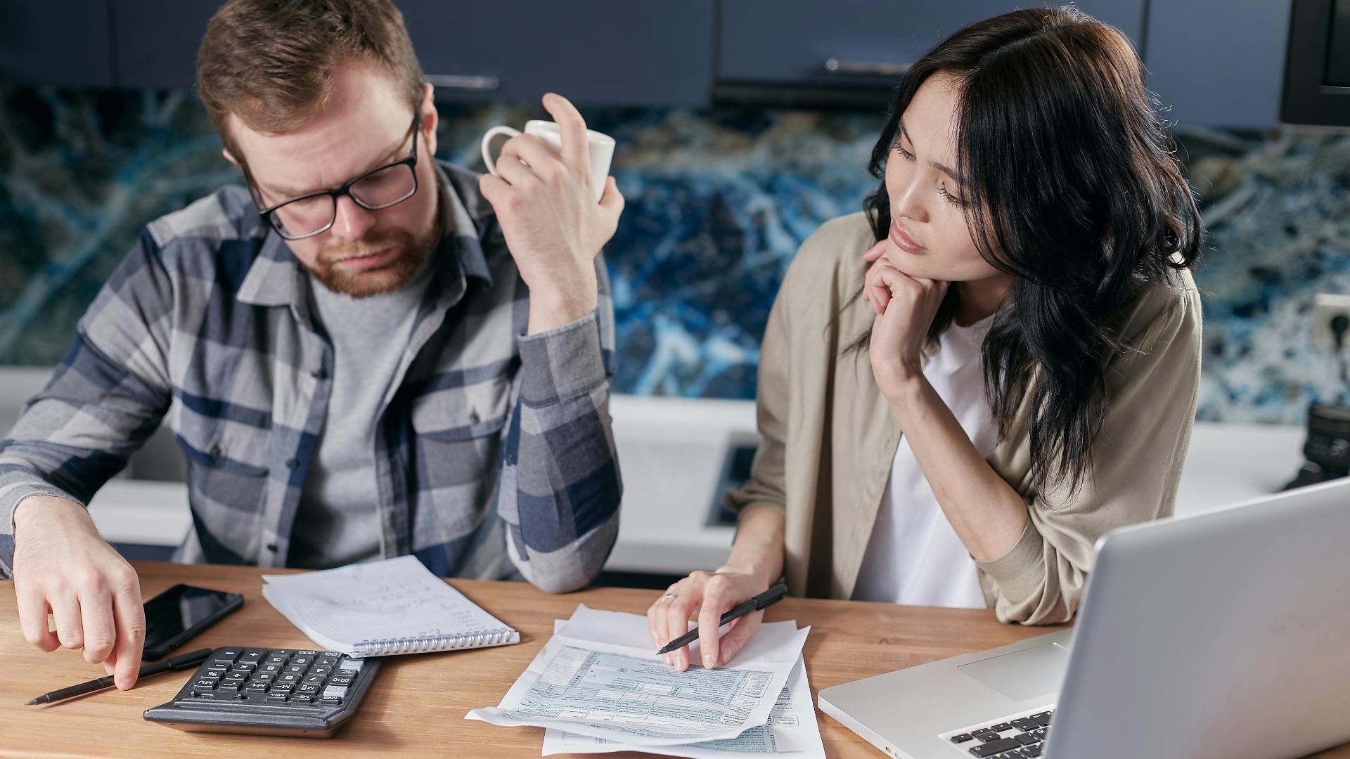 A couple reviews bills and documents on a laptop while discussing household finances.