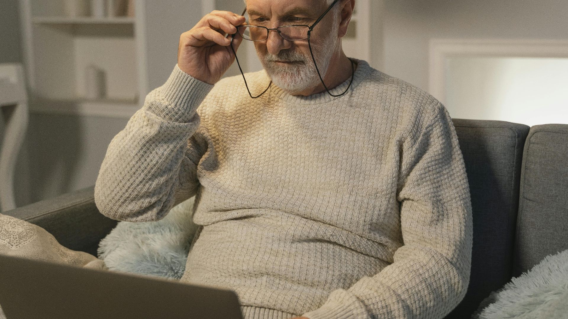 Elderly man wearing eyeglasses and a beige sweater, sitting on a sofa and using a laptop indoors.