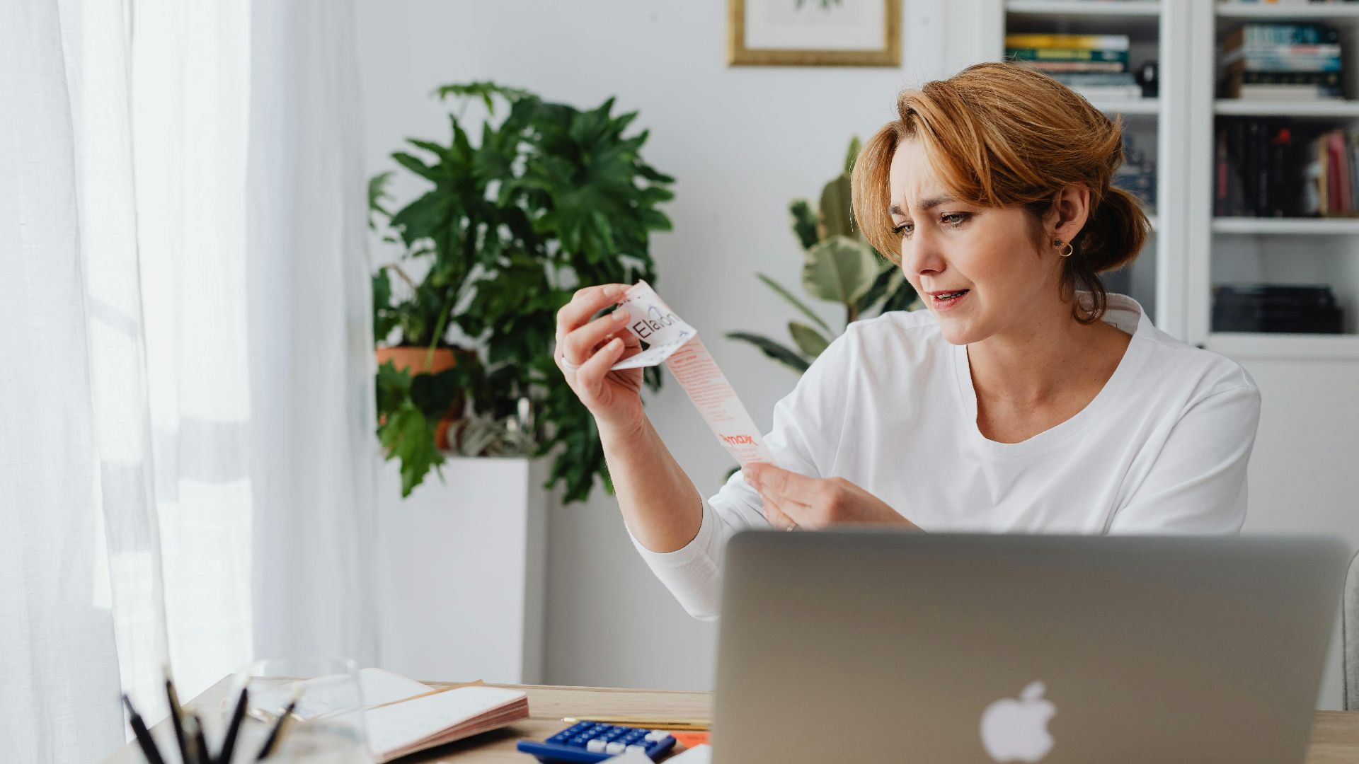 A woman in a white shirt sits indoors, examining a lengthy receipt with a concerned expression.