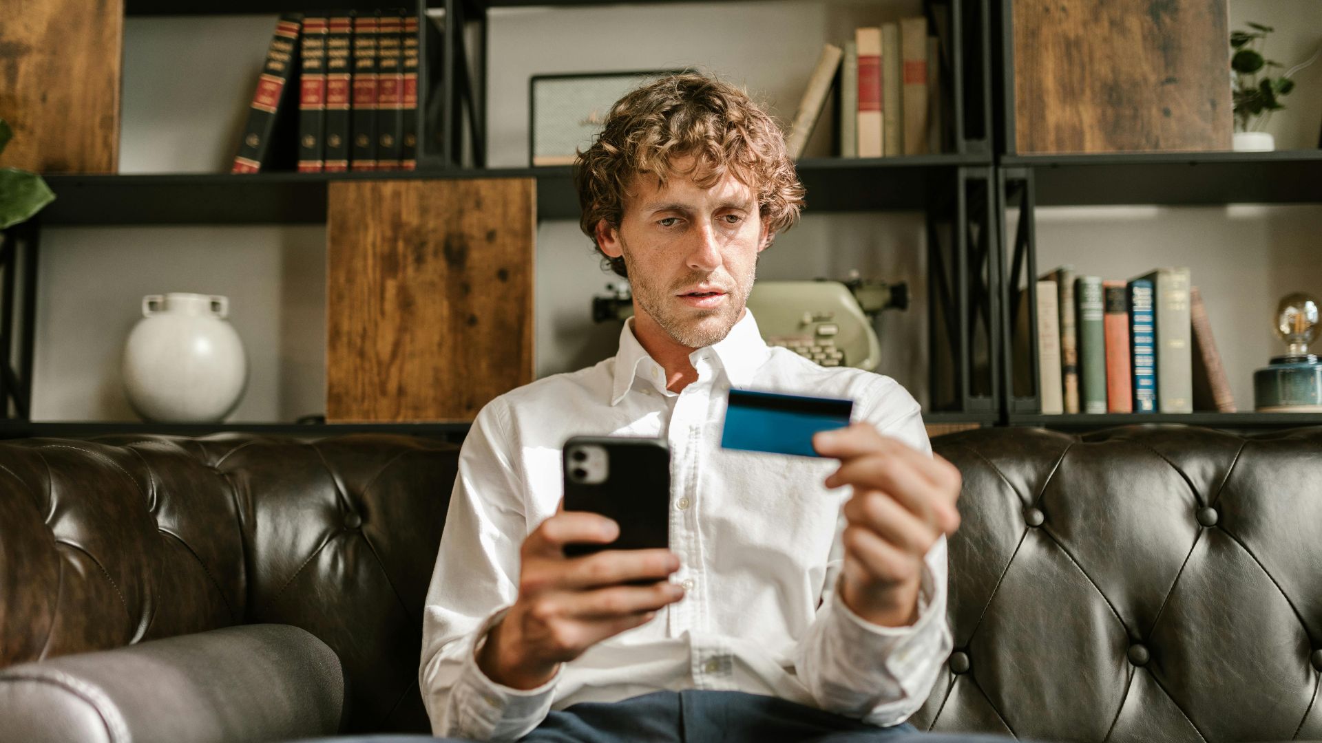 A man sitting on a leather sofa using a smartphone and credit card for online shopping.