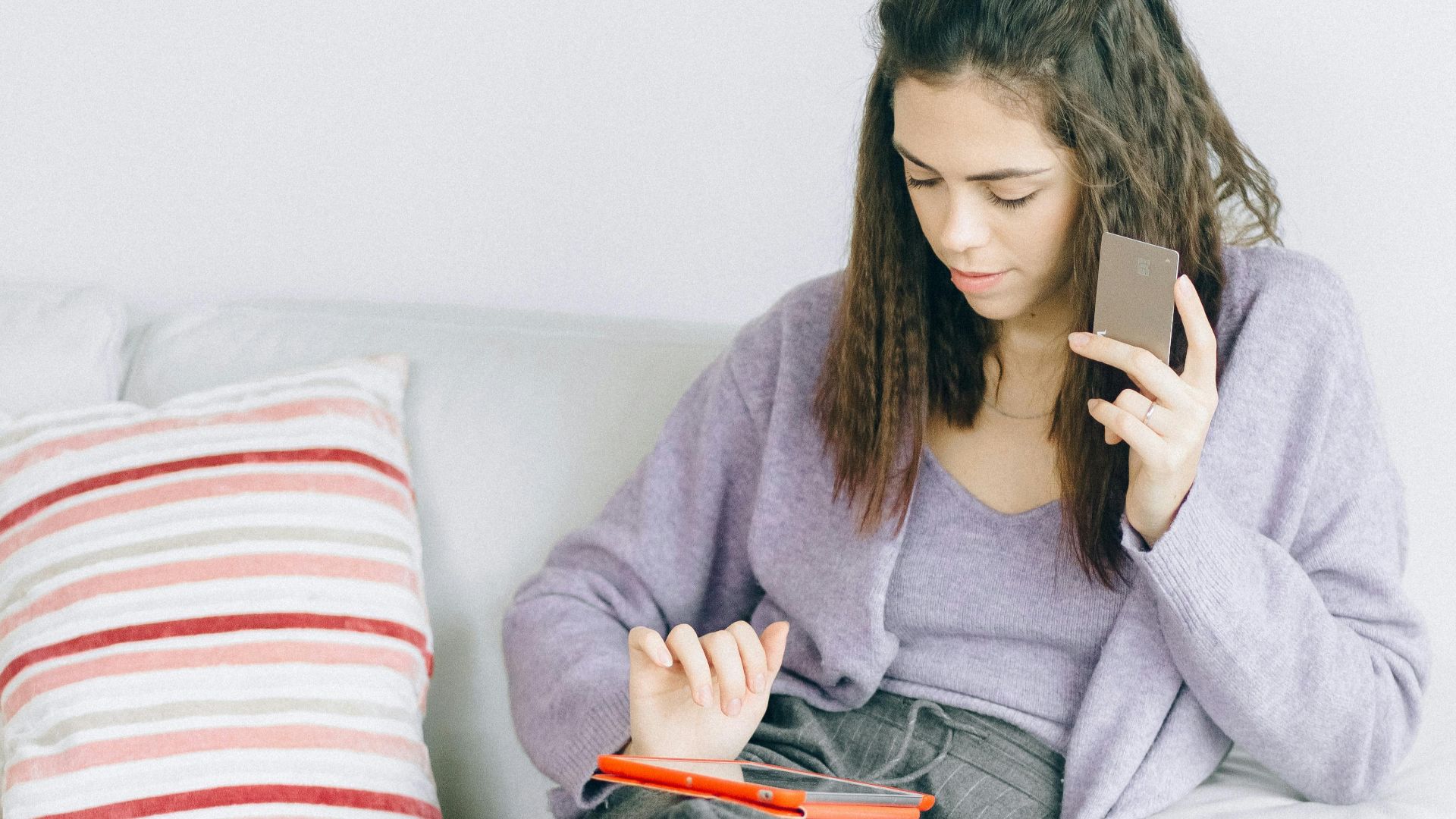 Woman sitting on a couch enjoying online shopping with a digital device and credit card.