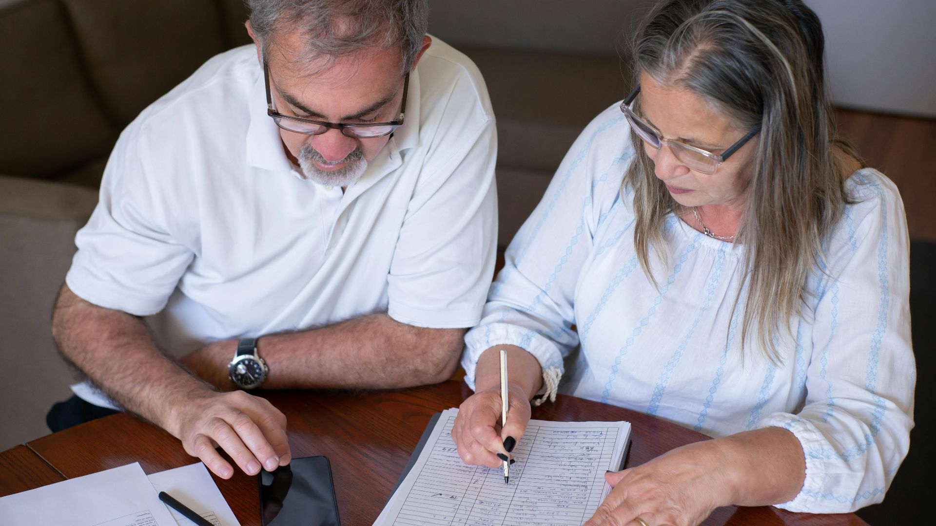 Elderly couple reviewing documents, using smartphone for online banking at home.