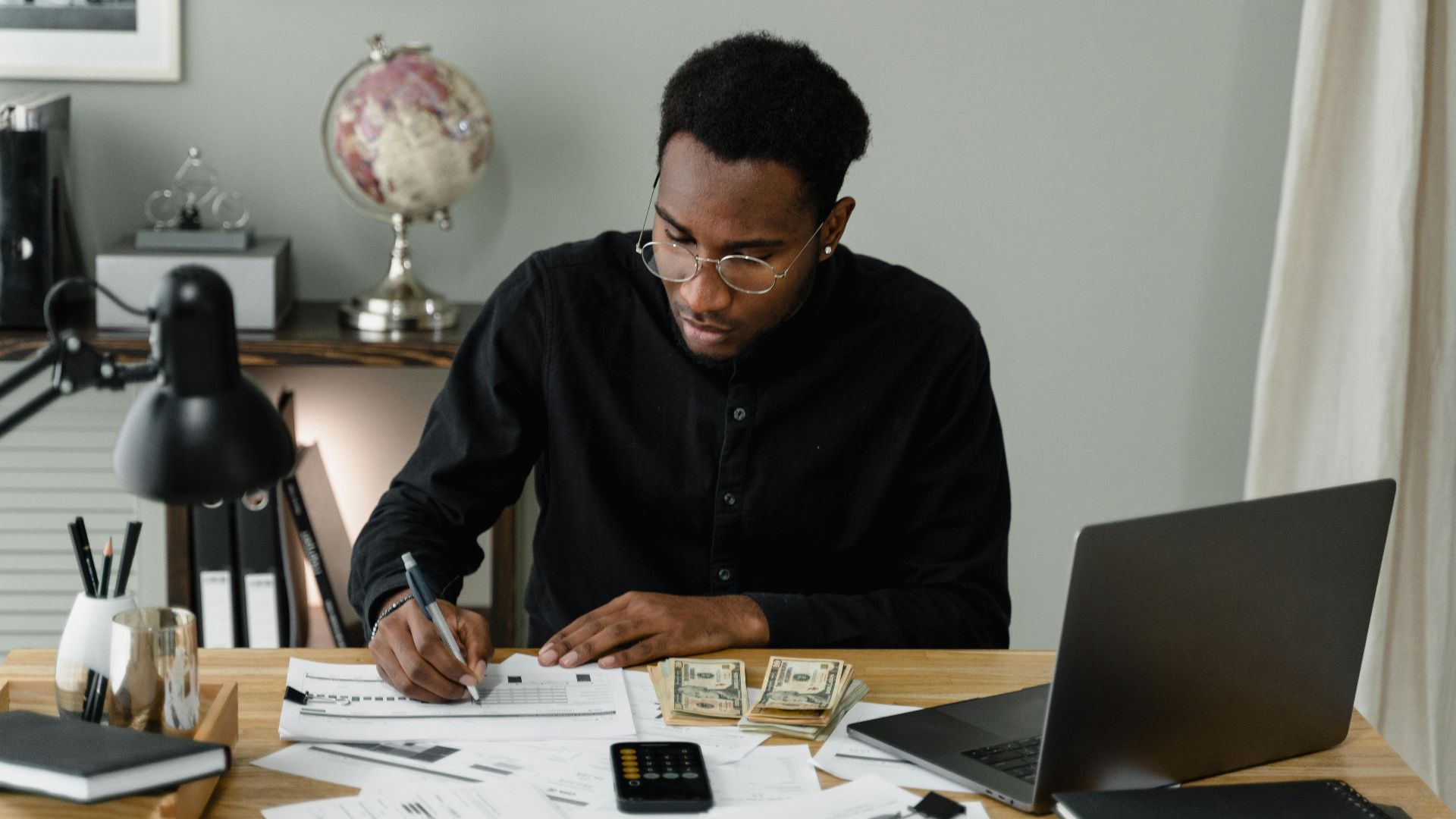 An adult man calculates expenses, using a laptop and documents at a desk in a home office setting.