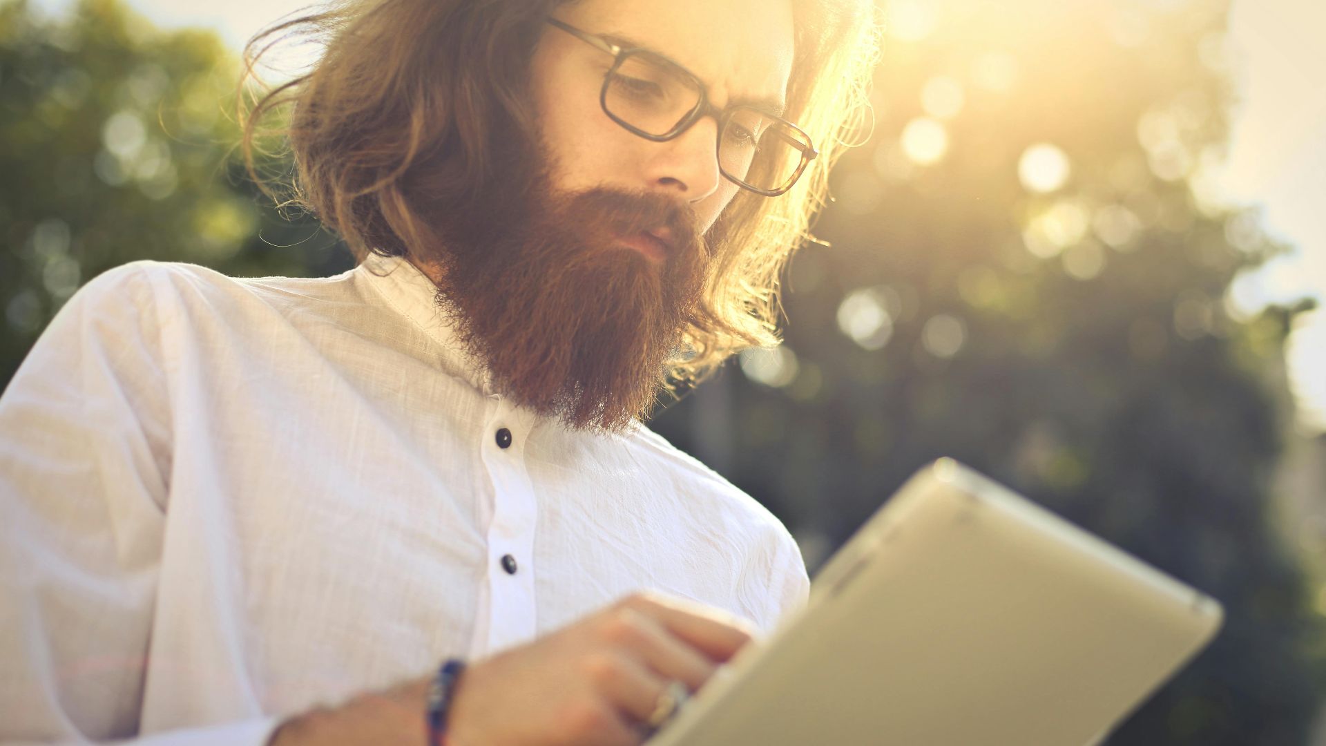 Bearded man using a digital tablet outdoors on a sunny day, focused and absorbed.