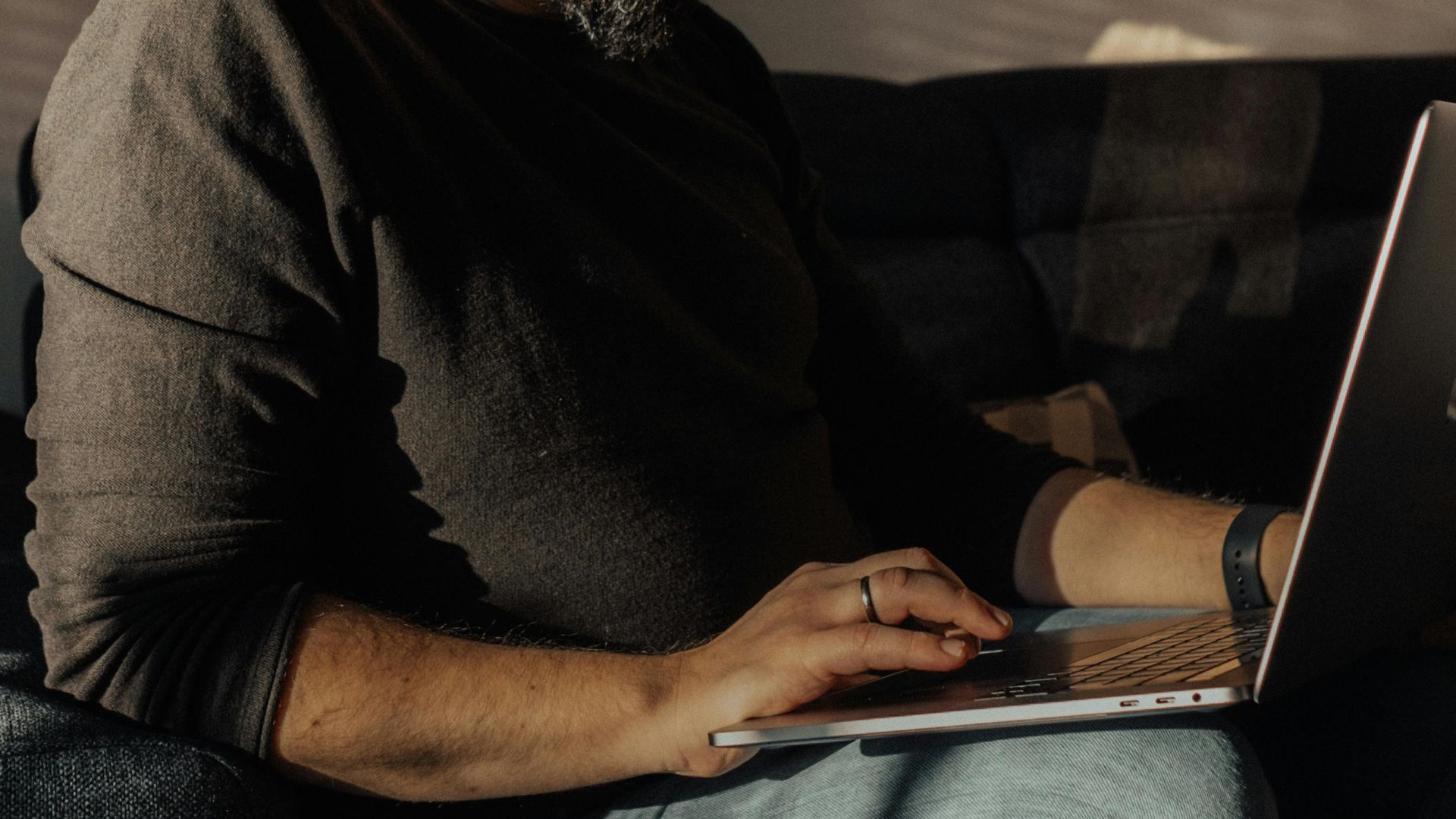 Man with beard and glasses using a laptop on sofa, sunlight streaming indoors.
