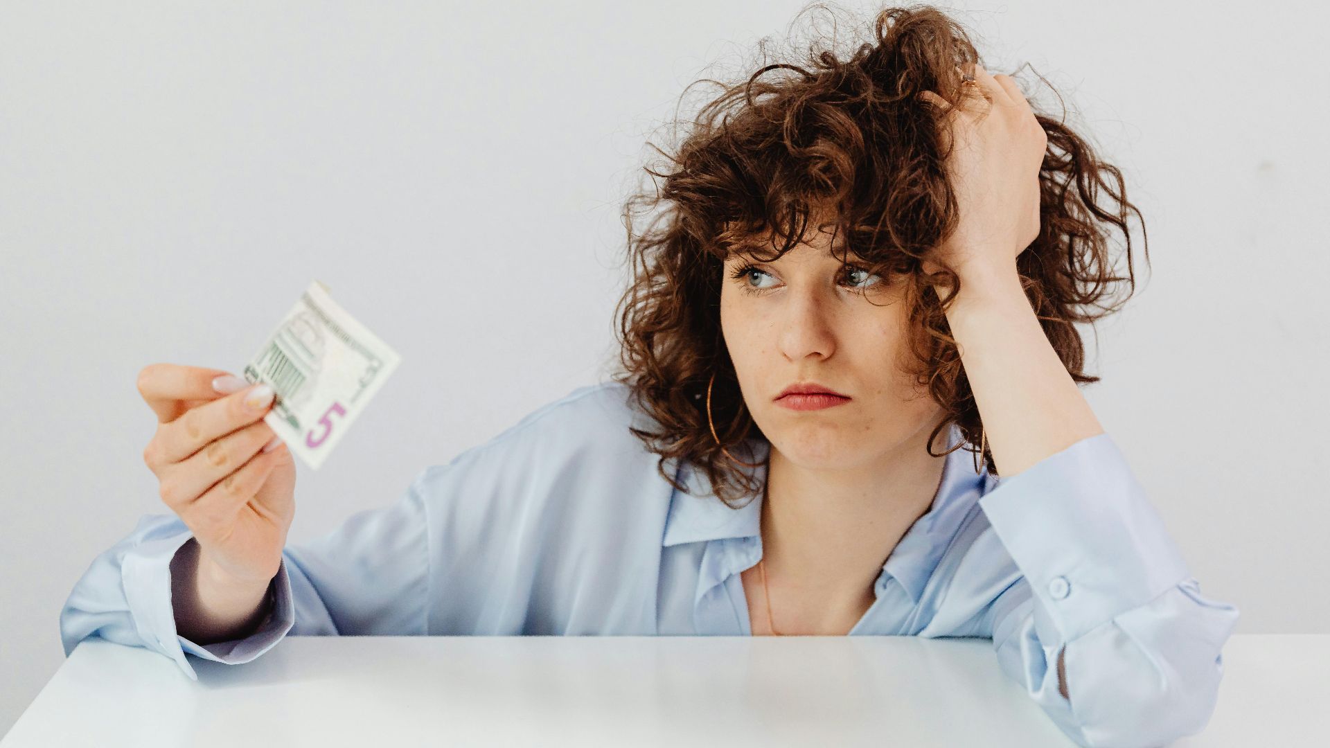 Concerned woman with curly hair holding a five dollar bill, expressing financial worry.